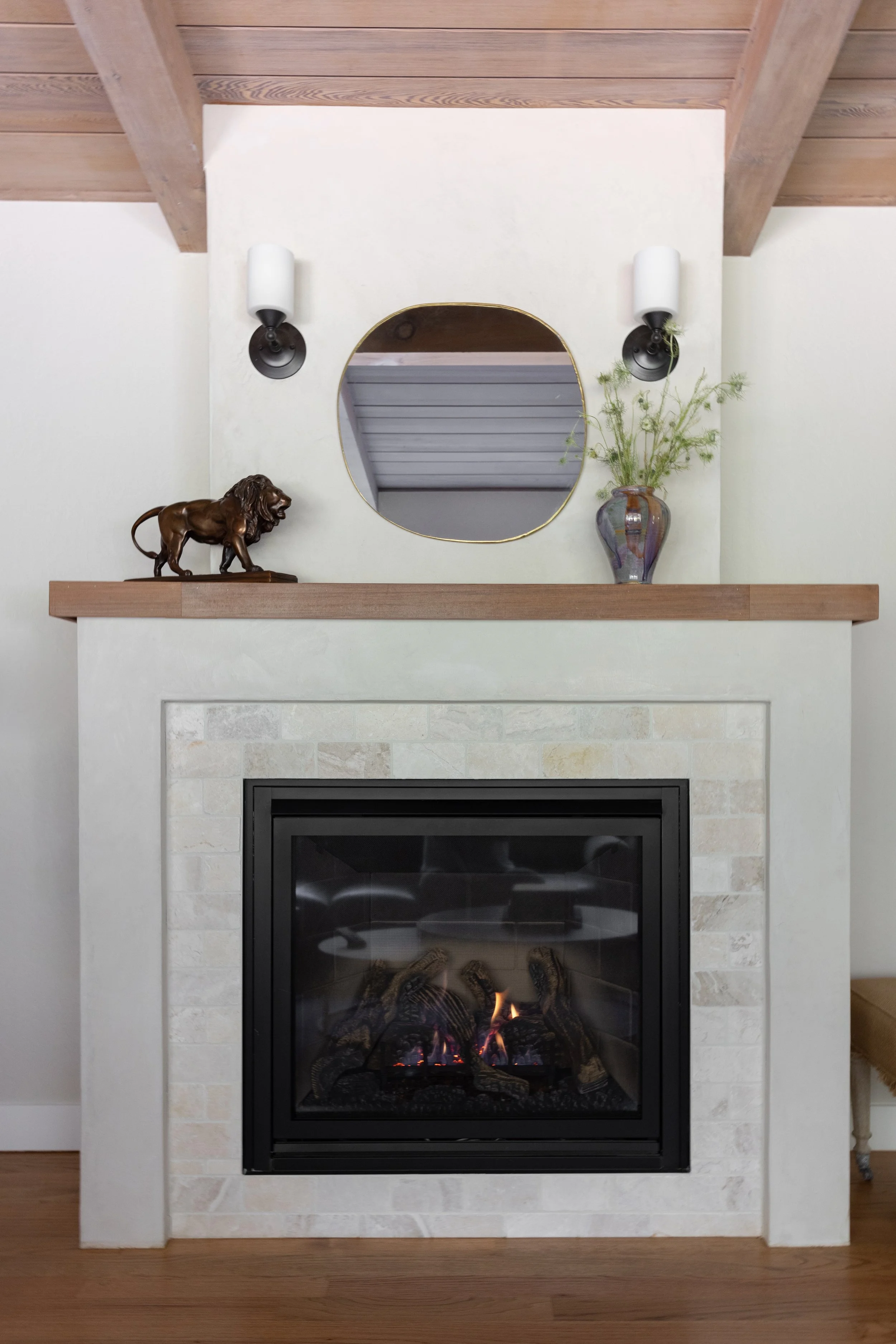 A fireplace with a white mantel, a glass door, and a fire burning inside. Above it is a round mirror, with a bronze lion figurine on the left and a vase with green foliage on the right. Two black and white wall sconces are mounted above.