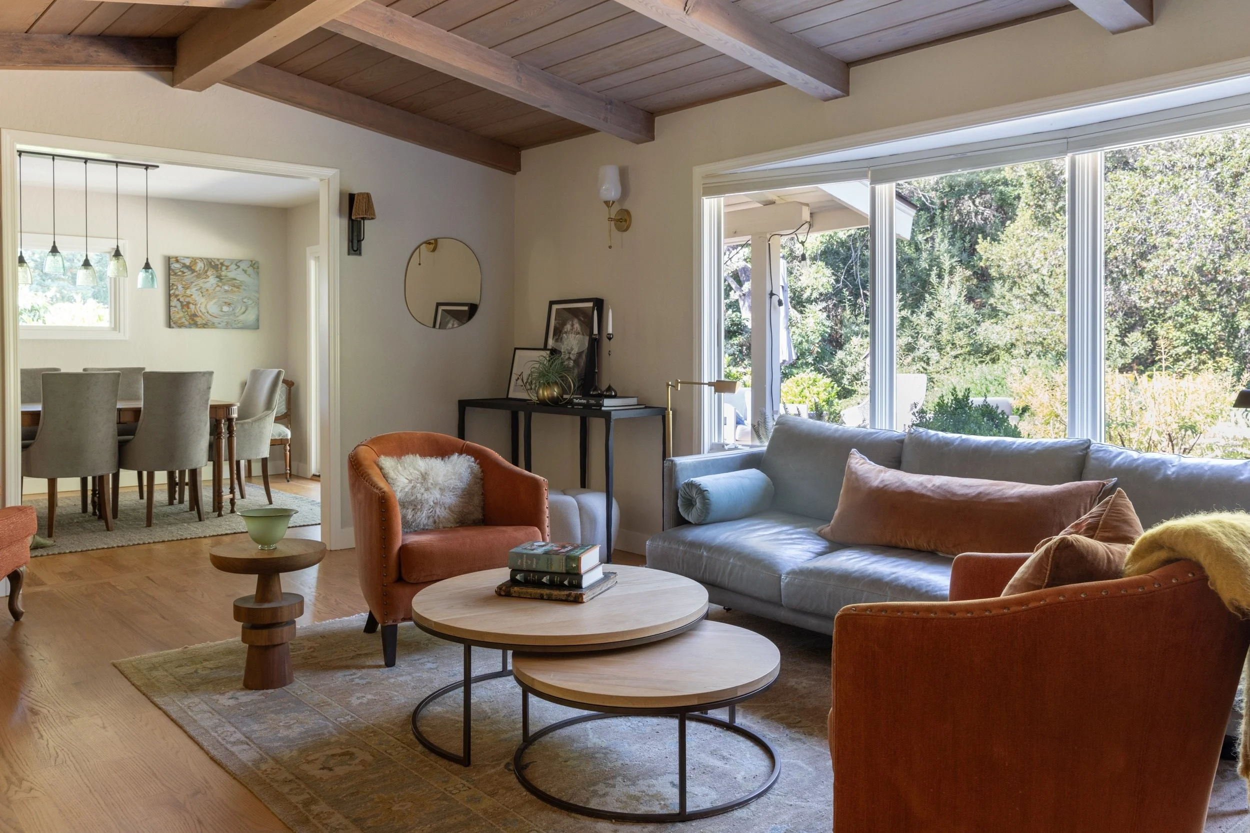 Living room with large windows, sofa, armchairs, wooden coffee tables, decor, and a view of the garden outside.