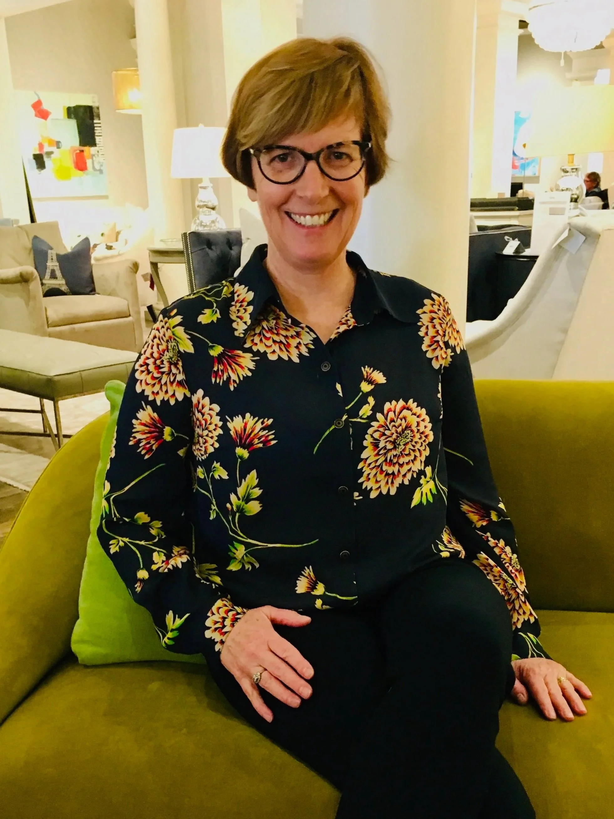 A smiling woman with short brown hair, glasses, wearing a black shirt with a colorful floral pattern, sitting on a yellow-green sofa in a furniture store or showroom.