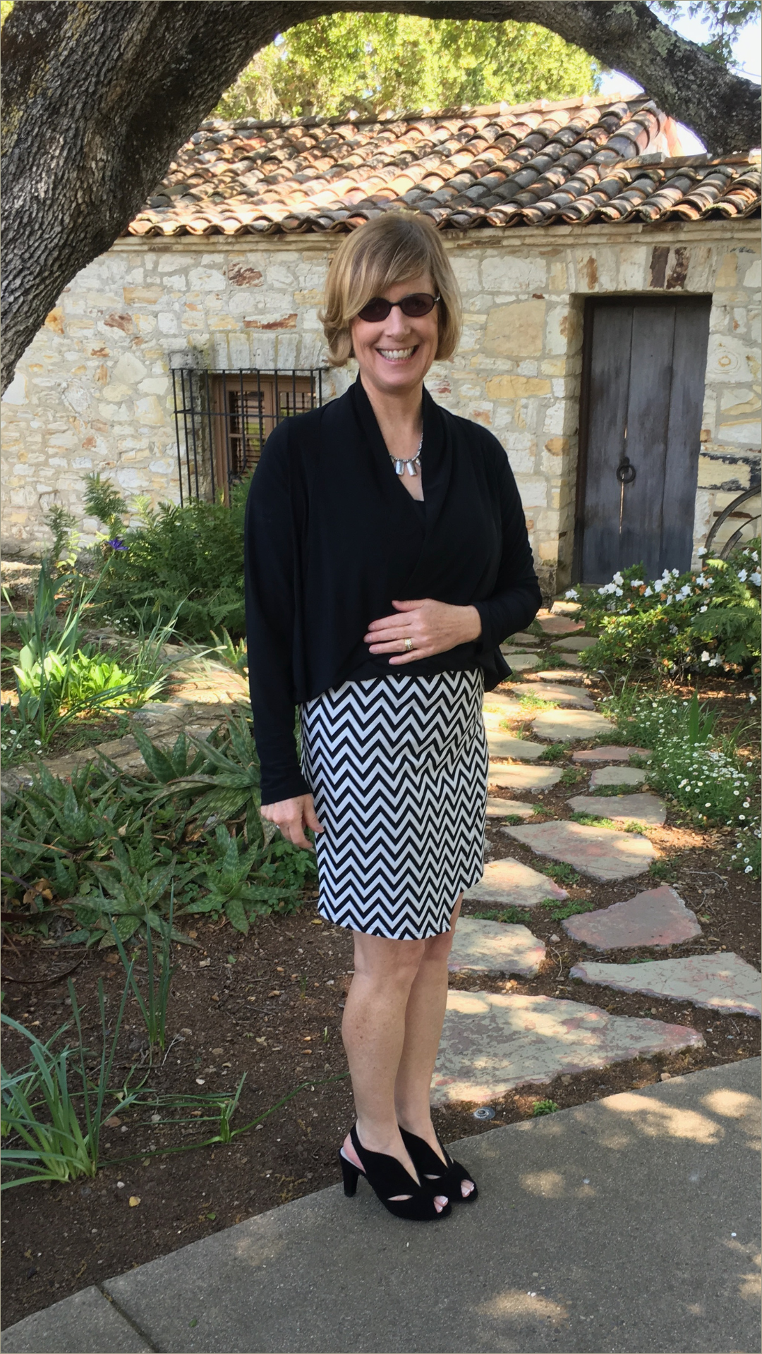 A smiling woman with short blonde hair, wearing sunglasses, a black jacket, a black and white chevron patterned skirt, and black high heel shoes, standing on a stone pathway in a garden under a large tree.