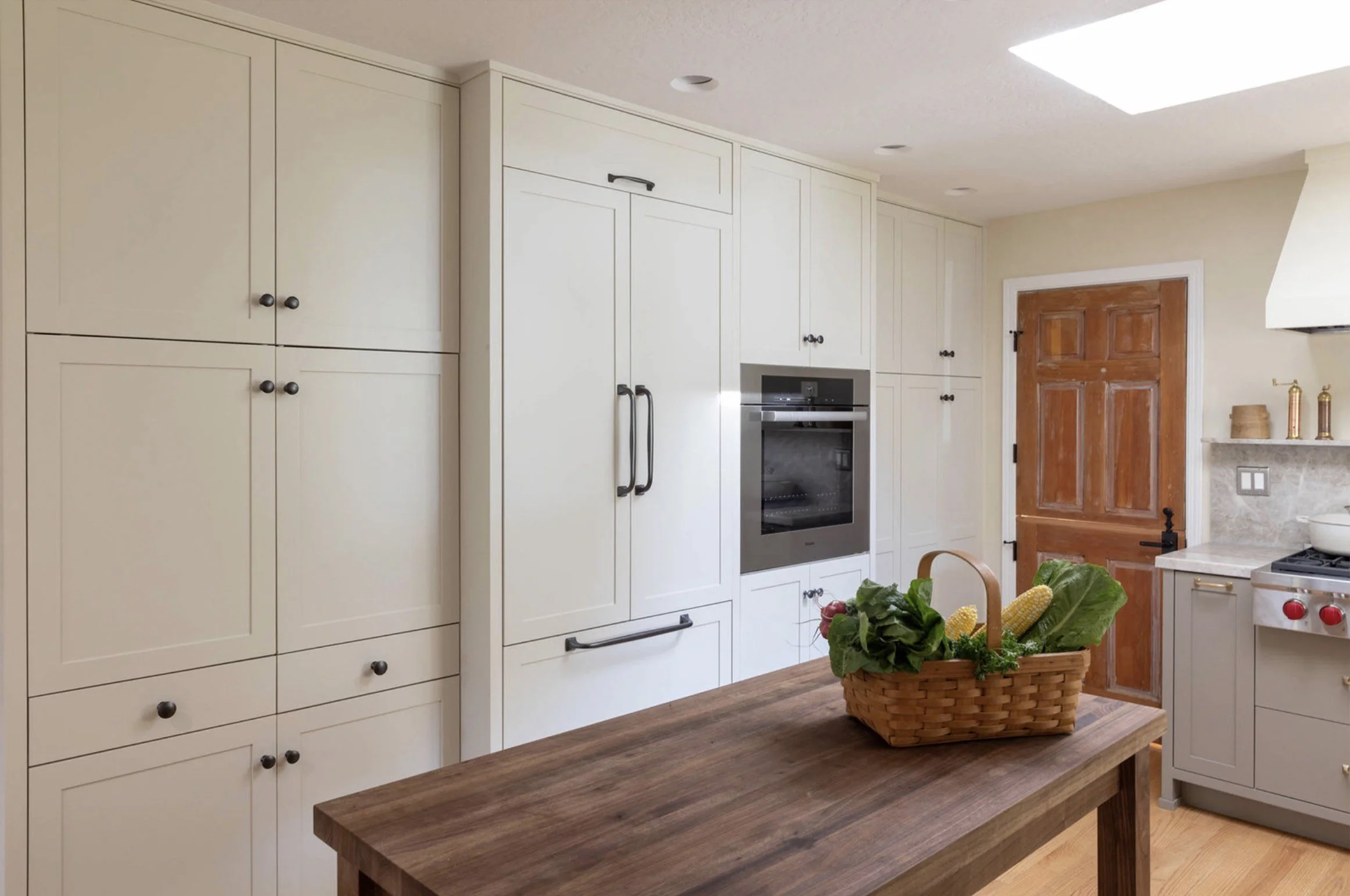 A kitchen with white cabinets, a built-in oven, a wooden door, a wooden table, and a basket with fresh vegetables.