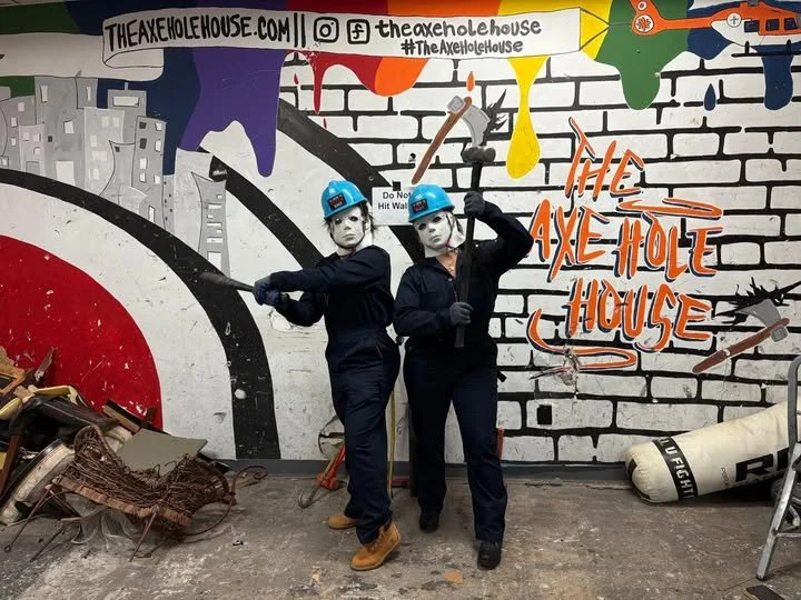 Two people wearing blue helmets and safety goggles stand in front of a graffiti wall at The Axe Hole House, with one holding an axe and the other posing with a bat. The wall features cityscape and colorful paint drips, and the text 'The Axe Hole Hous