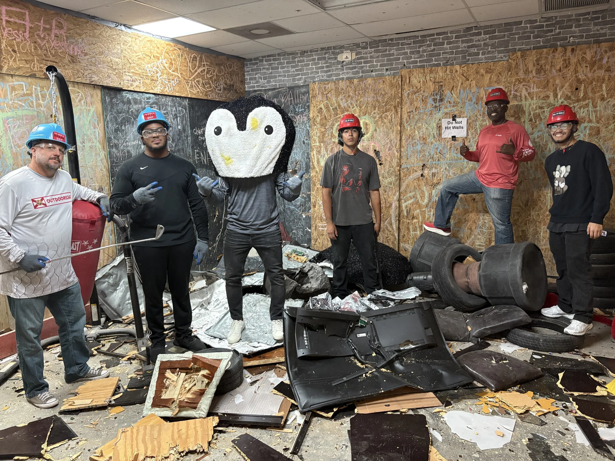 Group of five people in a damaged indoor space with debris and furniture, wearing safety gear including helmets and gloves. One person is dressed with a large penguin headpiece, and one is holding a sign that says "Do Not Hit Walls."