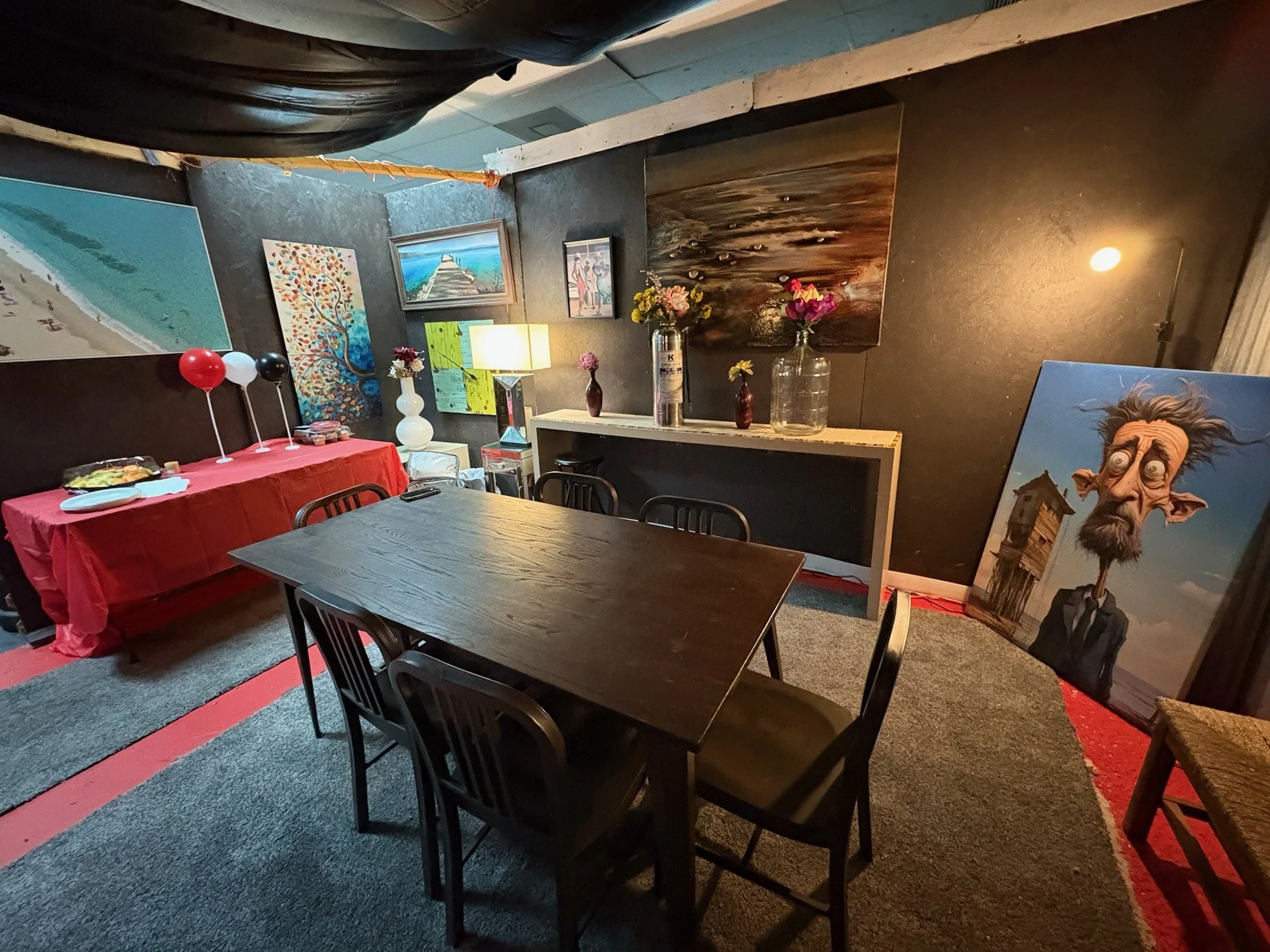 A decorated interior of a room with a wooden table and chairs, various paintings on dark walls, and a large caricature portrait leaning against the wall. There are vases with flowers on a white shelf, a lamp, and balloons on a red tablecloth in the corner.