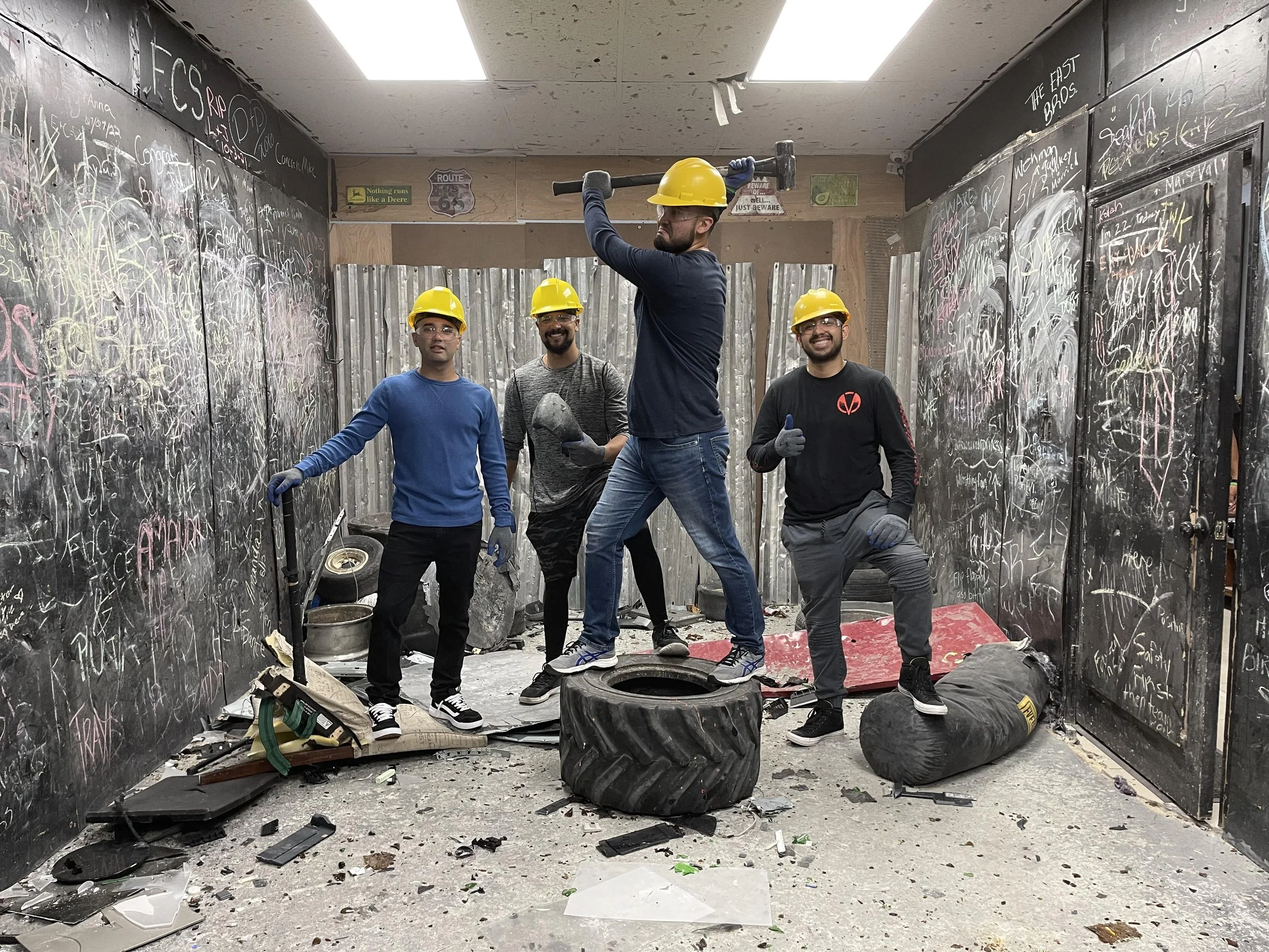 Four men wearing yellow hard hats and work gloves standing in a cluttered, graffiti-covered room with debris on the floor, a large tire, and various tools and objects.