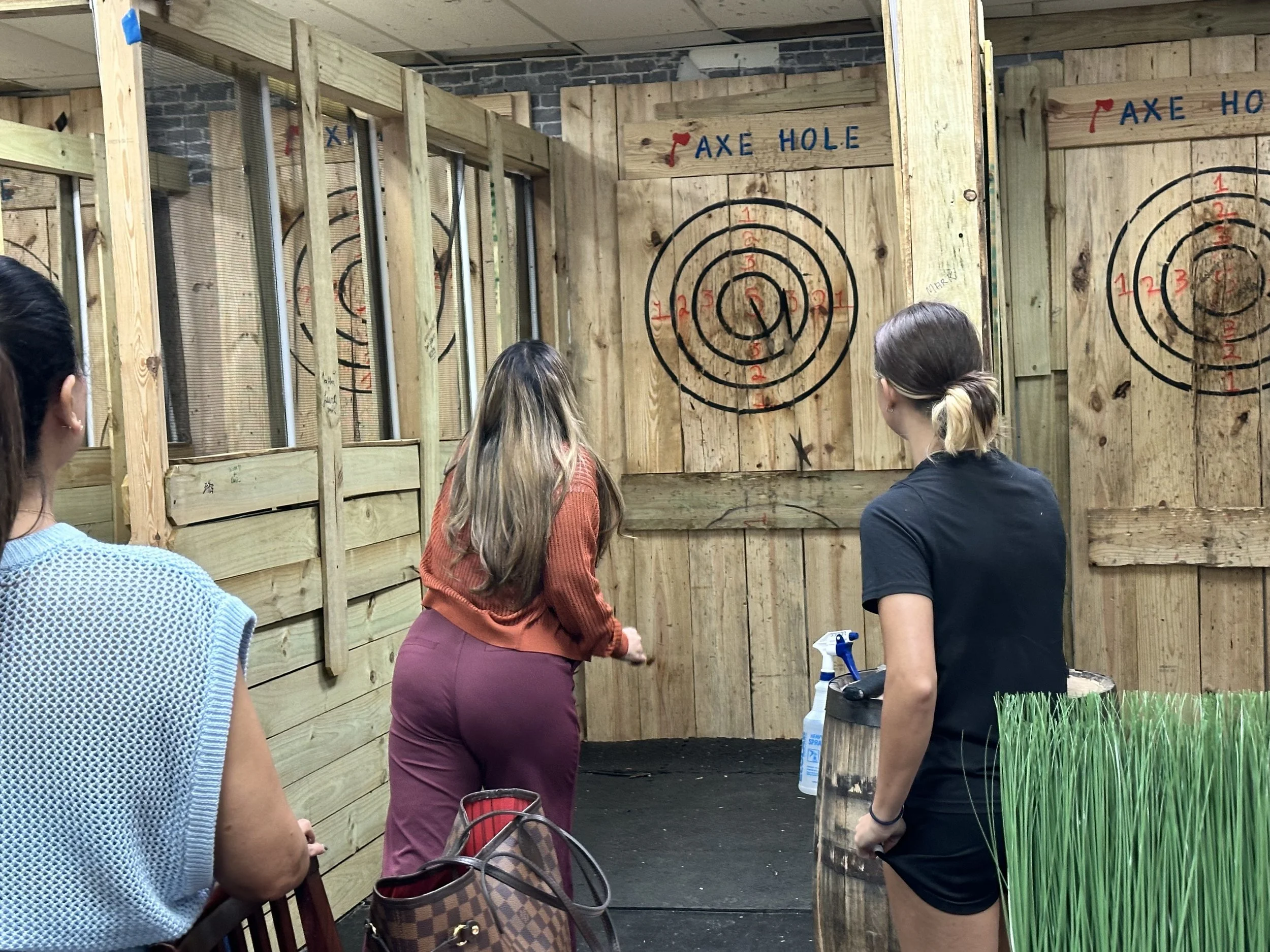 Three women playing axe throwing at an indoor axe-throwing venue with wooden targets on the wall.