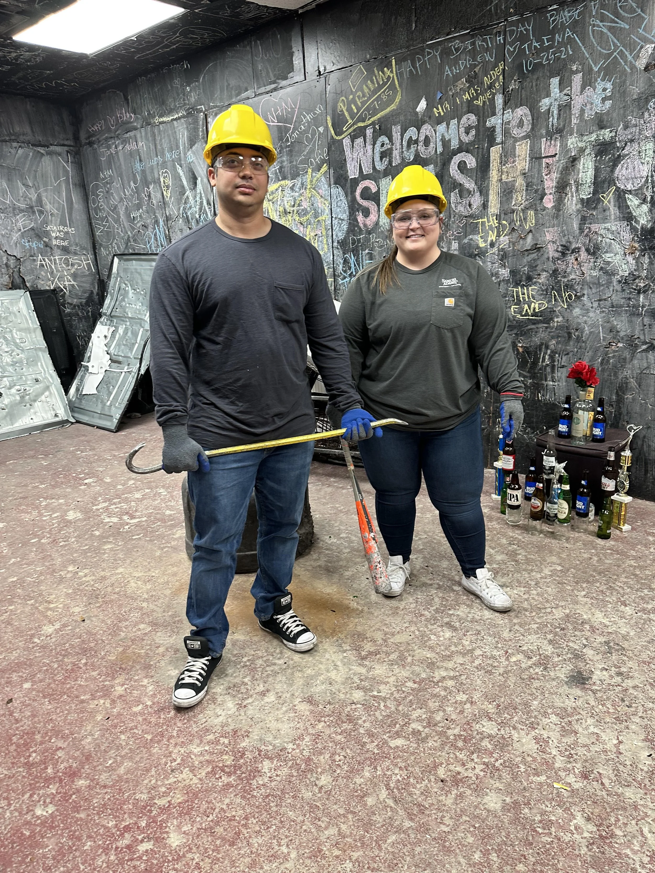 A man and a woman wearing yellow safety helmets and glasses, standing inside a graffiti-covered room, holding a crowbar and a hammer. The room has a black chalkboard wall with colorful chalk messages and a small table with multiple bottles and a flow