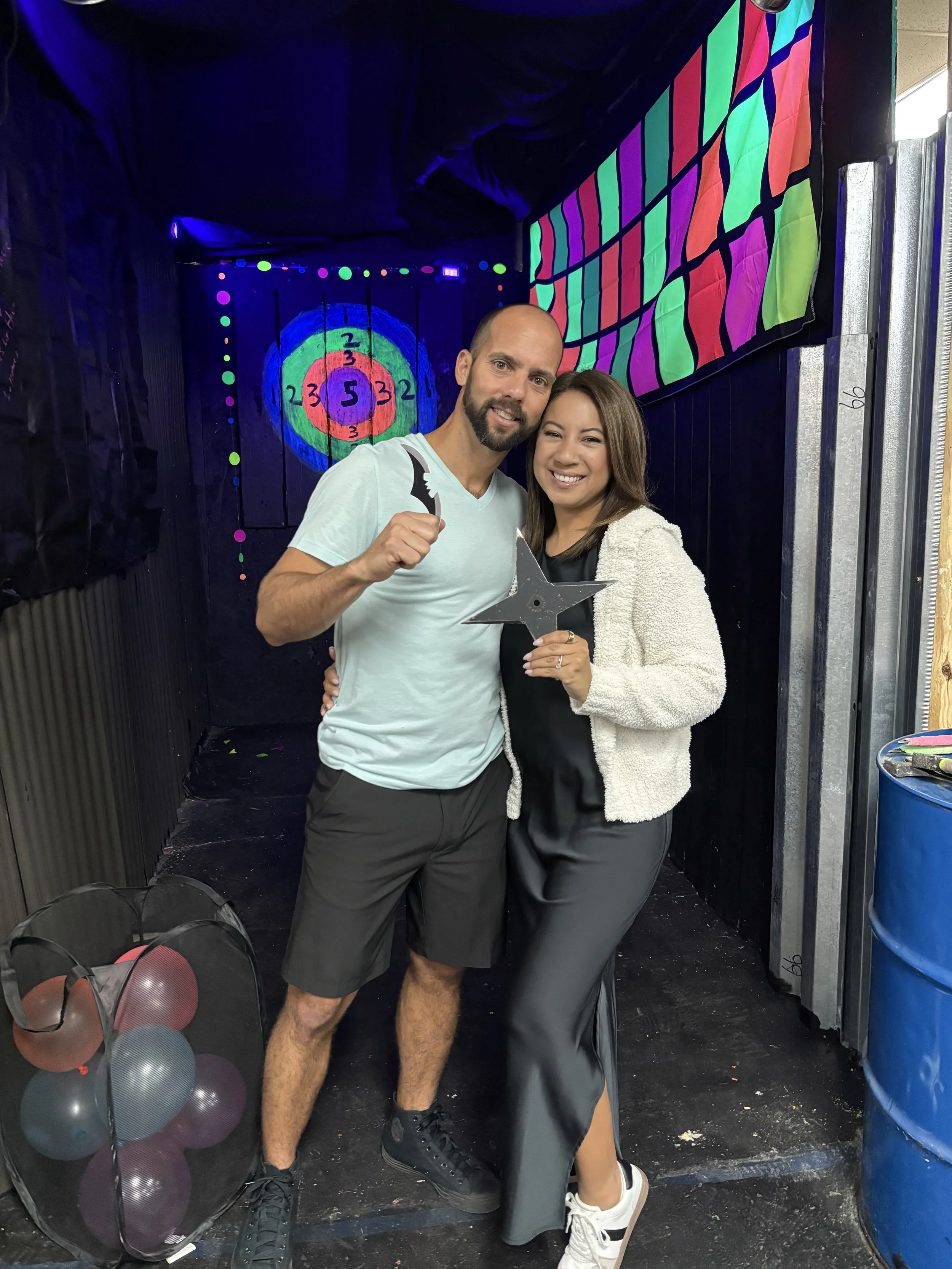A man and woman smiling and posing together in a room with a colorful background. The man is holding a small bat-shaped object and making a fist gesture. The woman is holding a star-shaped object. There are balloons in a black net bag on the floor, and a target with concentric circles is painted on the wall behind them.