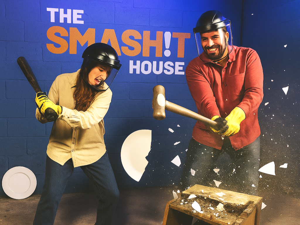 Two people wearing helmets and gloves smashing a wooden table with hammers inside a room with a blue wall and a sign that says "The Smash House."