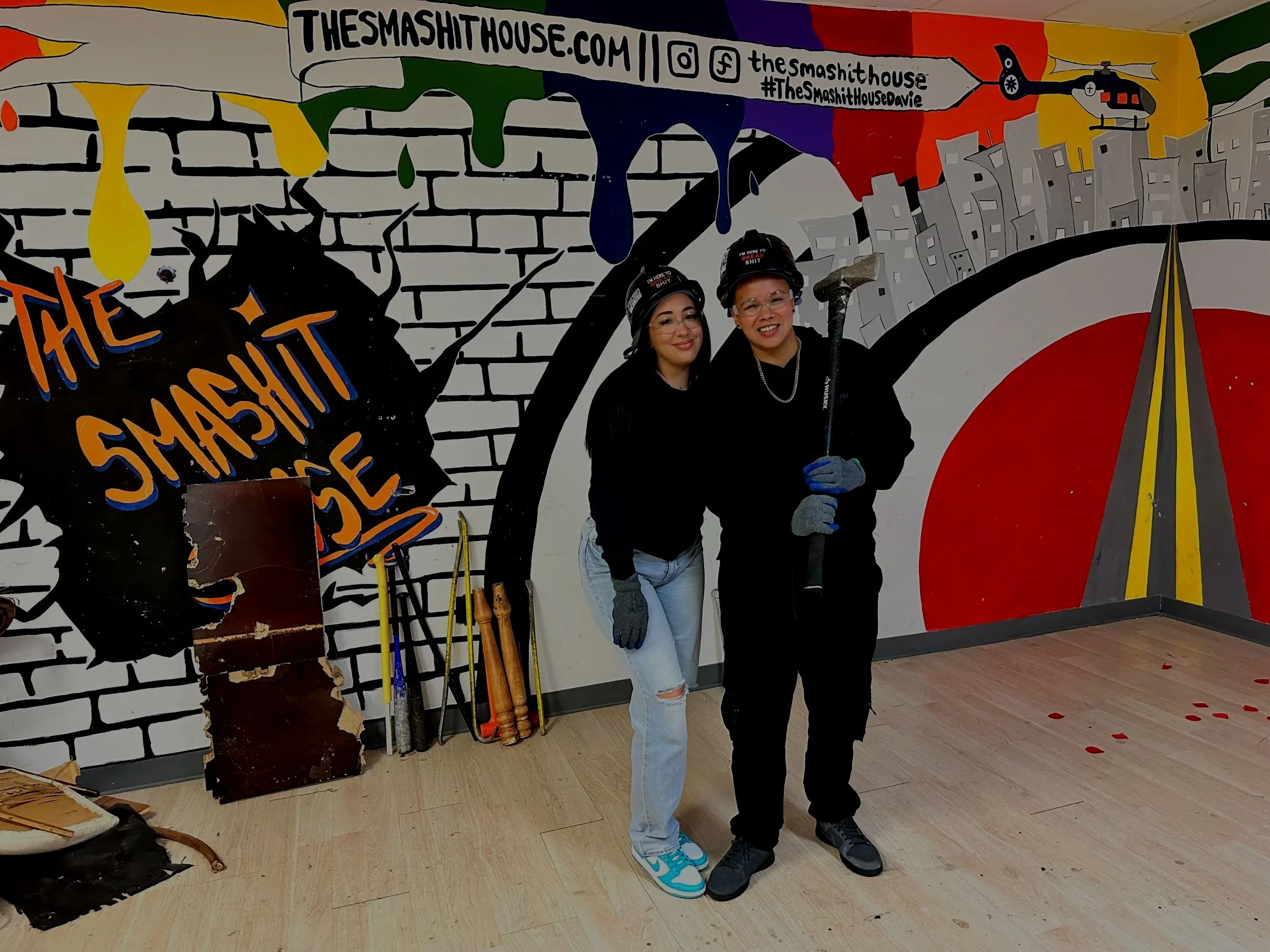 Two women wearing black helmets with the text "I'm here to break shit" are posing in front of a colorful mural featuring a helicopter, a cityscape, a brick wall, and abstract shapes. One woman is wearing a black jacket and glasses, and the other is wearing a black hoodie and glasses.