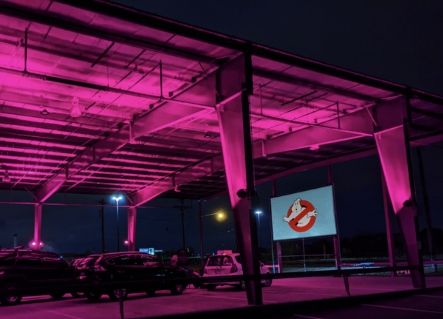 A parking lot under a roof with pink neon lighting and a large screen displaying the Ghostbusters logo at night. The location of GLIMMER, the pop-up drive-in created by Kris Growcott.