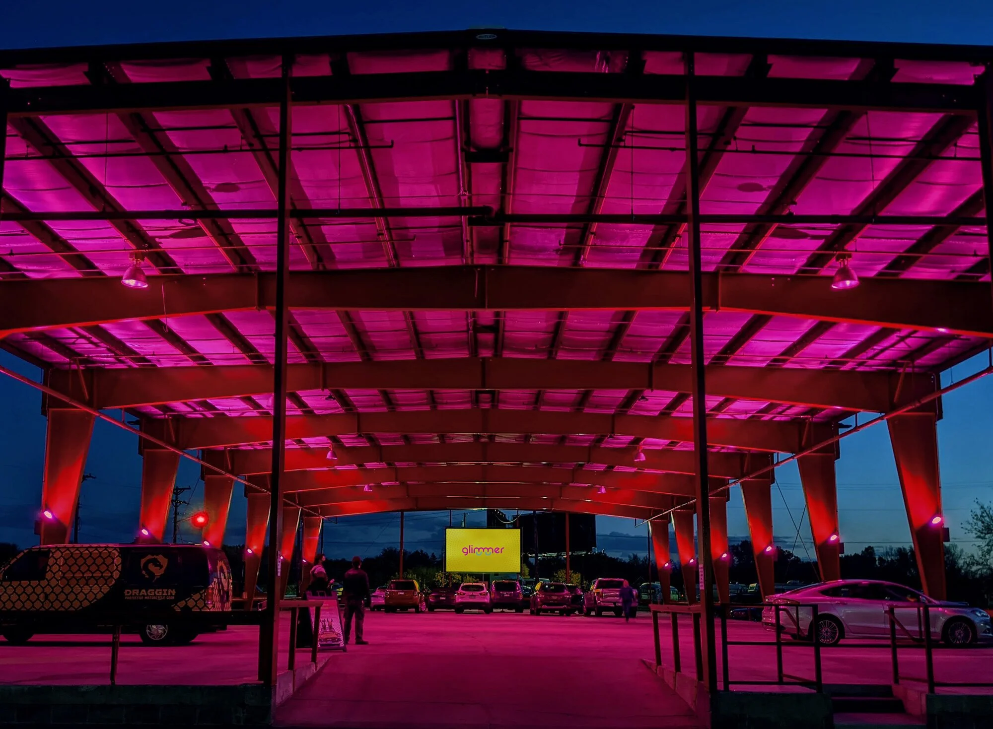 Outdoor parking area at night with a large, modern canopy lit with pink and purple lights, several parked cars, and people near a digital sign that reads 'glimmer'. The location of GLIMMER, the pop-up drive-in created by Kris Growcott