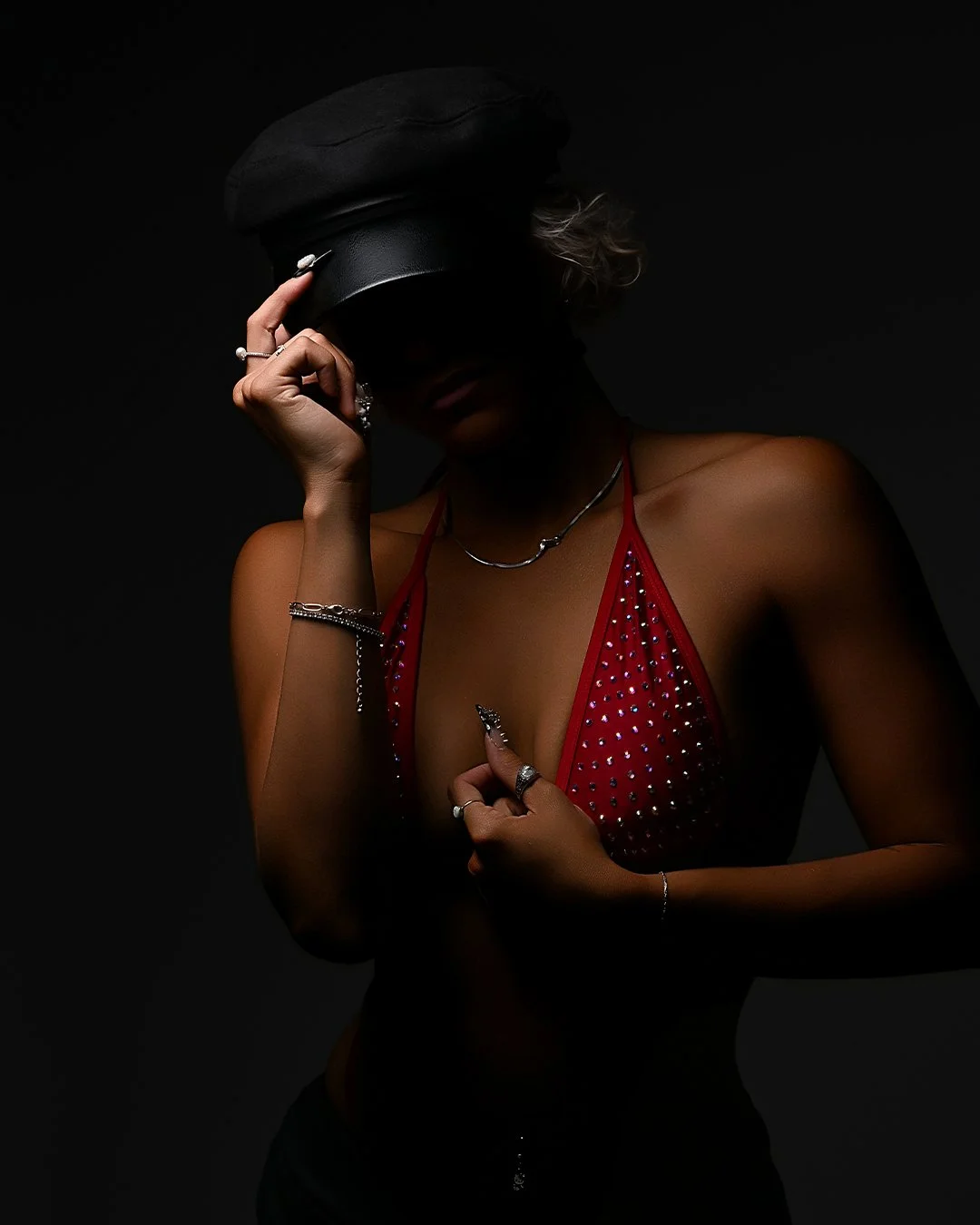 A woman wearing a black cap and a red rhinestone-studded top, holding the brim of her cap with one hand, with jewelry on her fingers, wrist, and neck, against a dark background.