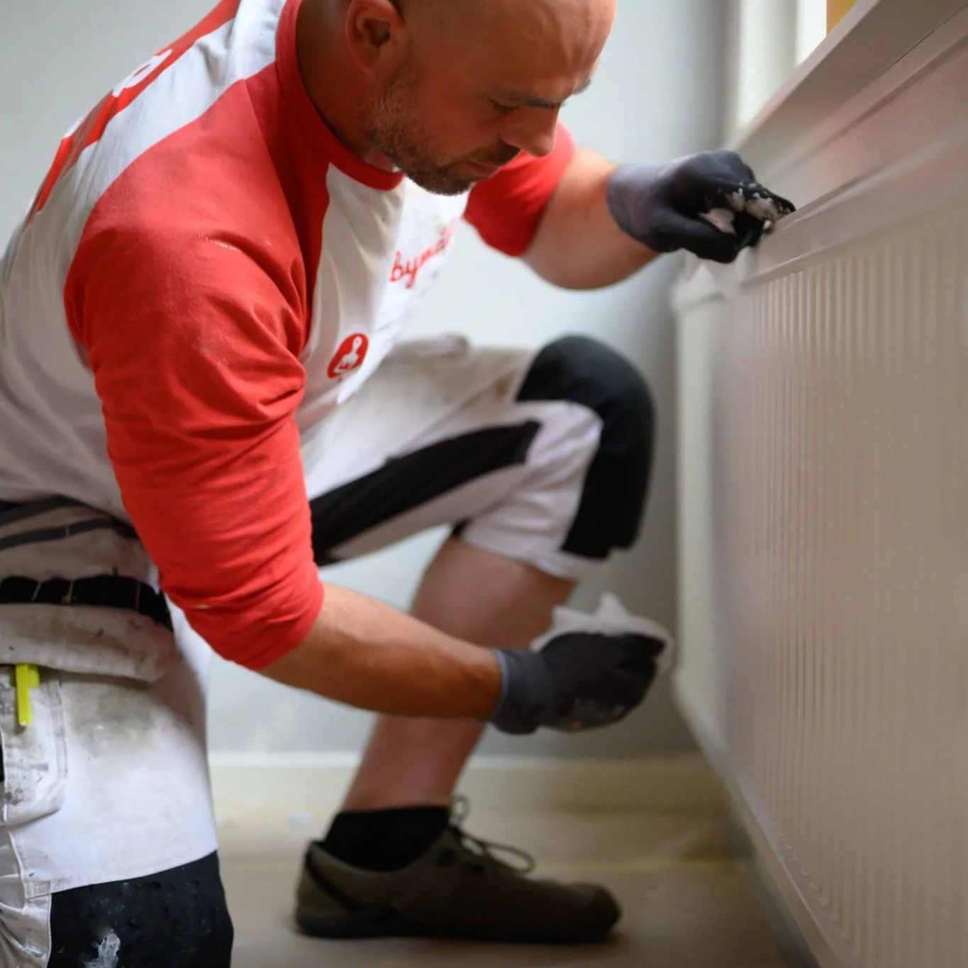 En man reparerar en radiator, klädd i arbetskläder och skyddshandskar, på knä vid en vägg.