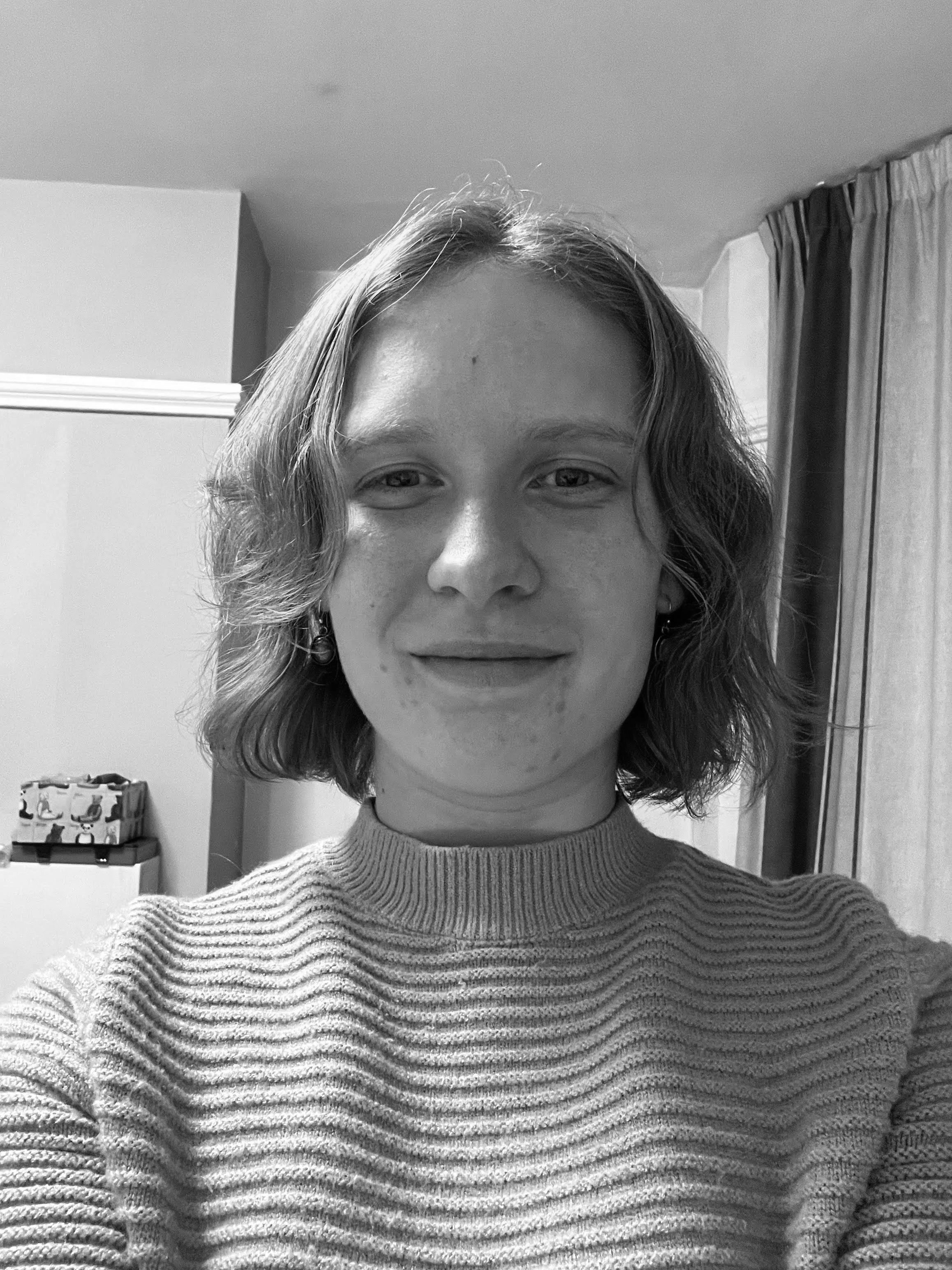 Close-up black and white photo of a young woman with shoulder-length wavy hair, wearing a ribbed sweater and earrings, smiling slightly, indoors with curtains and a dresser in the background.