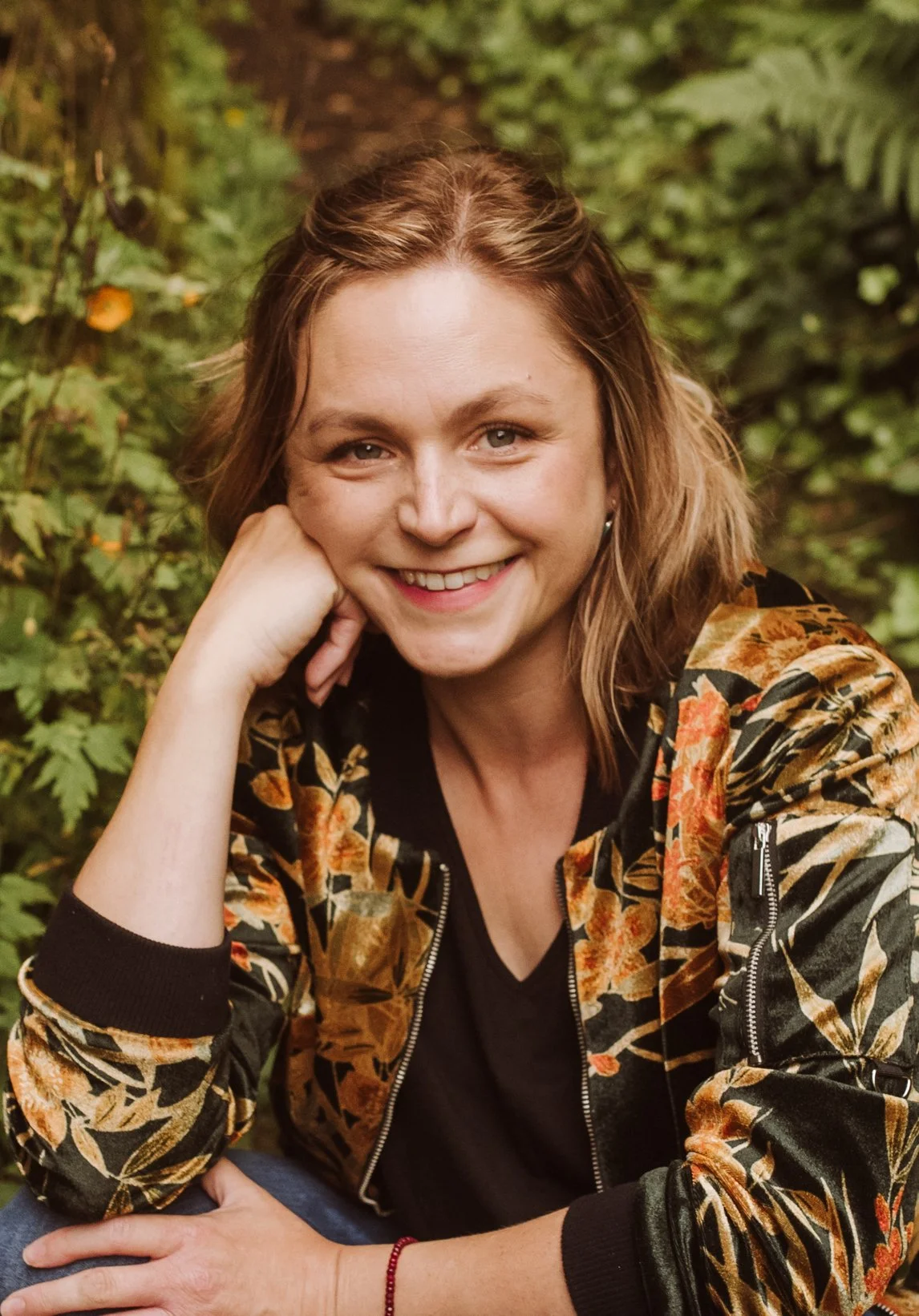 A smiling woman with light brown hair in loose waves, wearing a black shirt and a floral bomber jacket, sitting outdoors with green foliage in the background.