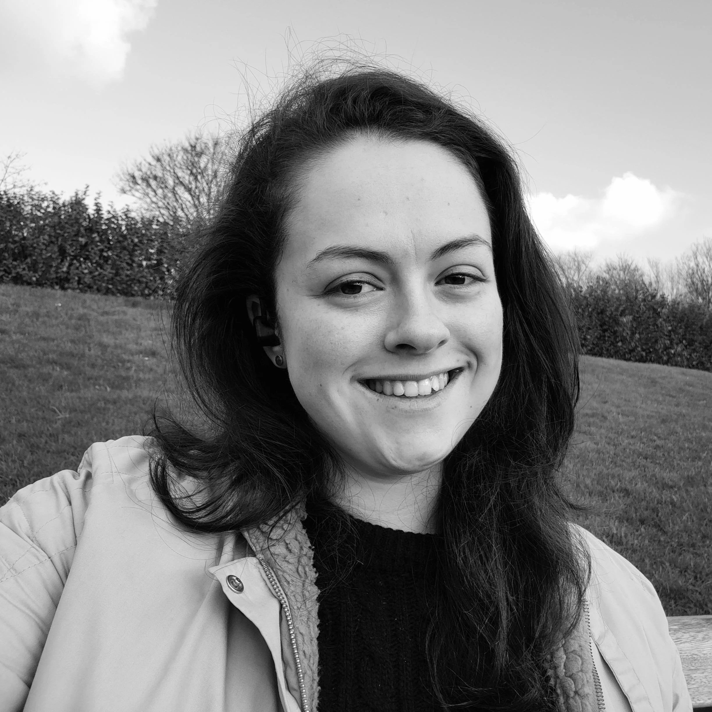 Black and white outdoor portrait of a young woman smiling, wearing a jacket, with trees and grass in the background.
