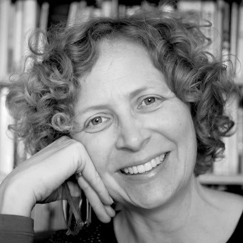 Alice Willitts, a woman with curly hair smiling and resting her head on her hand in front of a bookshelf.