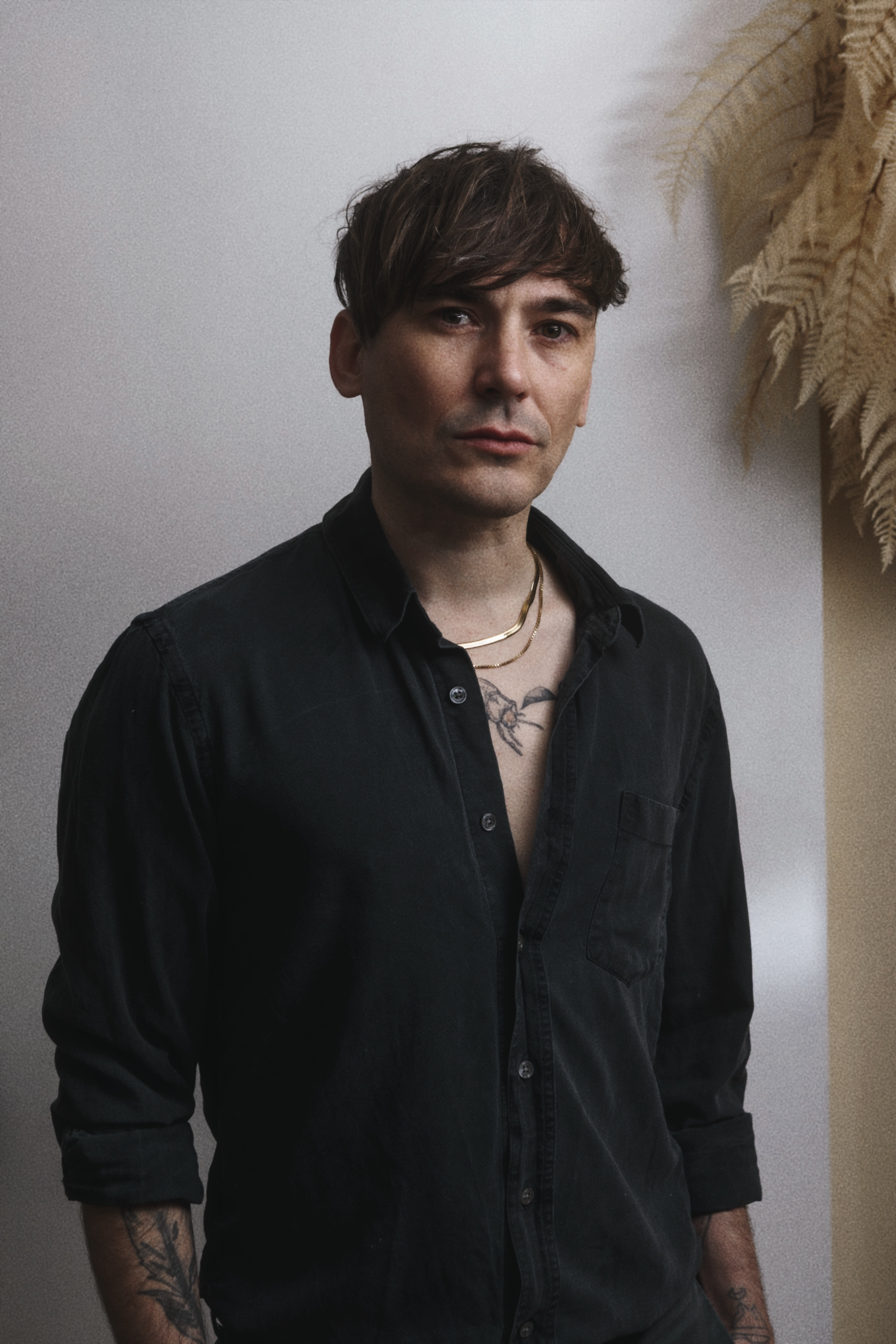 Andrew Hykel Mears with dark brown hair, wearing a black shirt with the top buttons open, revealing a tattoo on his chest and gold necklaces. He is standing indoors near a white wall and some dried plants in the background.