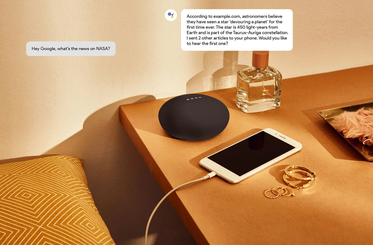 A wooden bedside table with a black smart speaker, a white smartphone connected to a charger, a transparent perfume bottle with a wooden cap, a tray with pink fuzzy fabric, and some gold jewelry including rings and bracelets. A pillow with an orange 