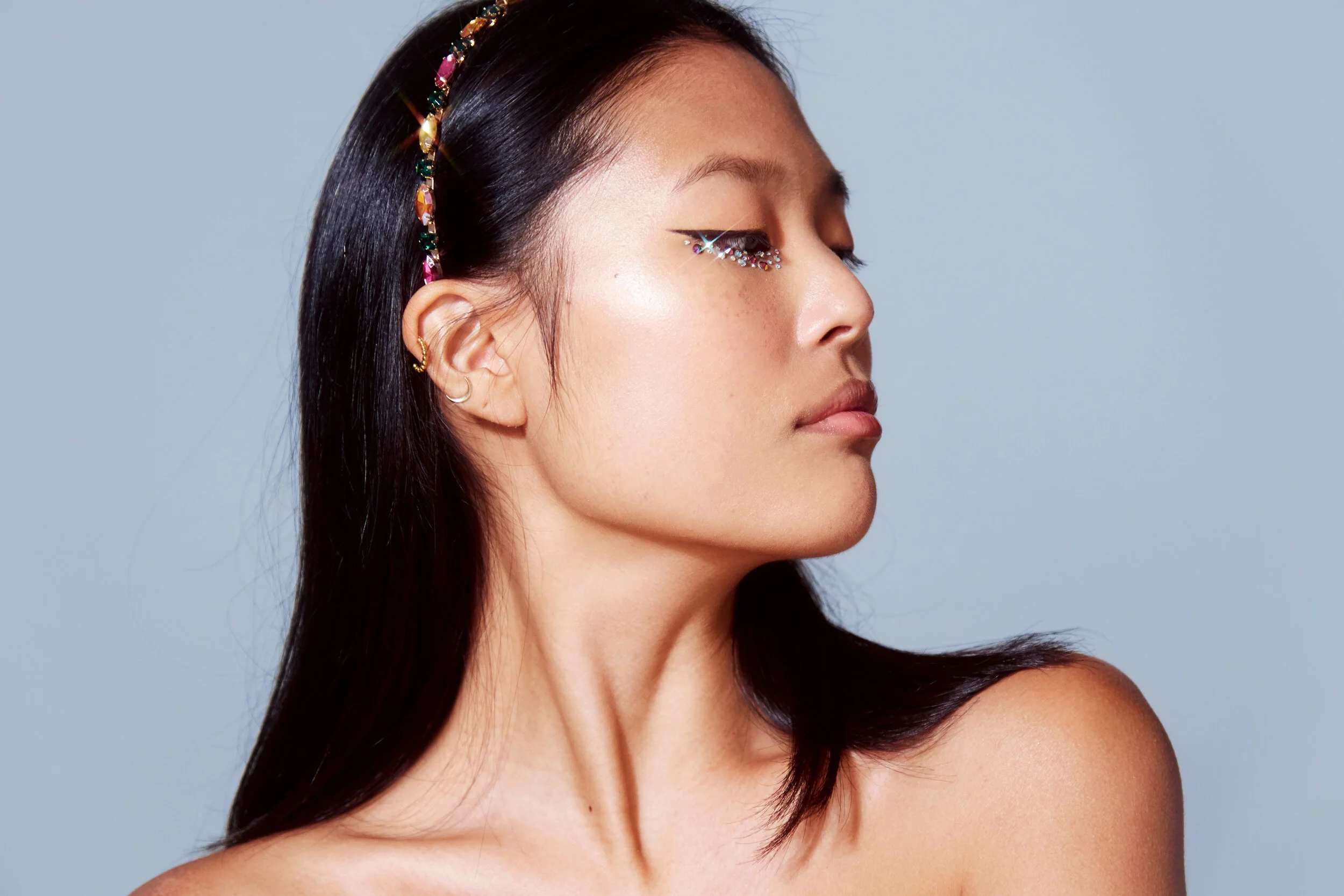 Close-up side profile of a woman with long dark hair, wearing a colorful beaded headband and sparkling rhinestone eye makeup, against a plain light blue background.