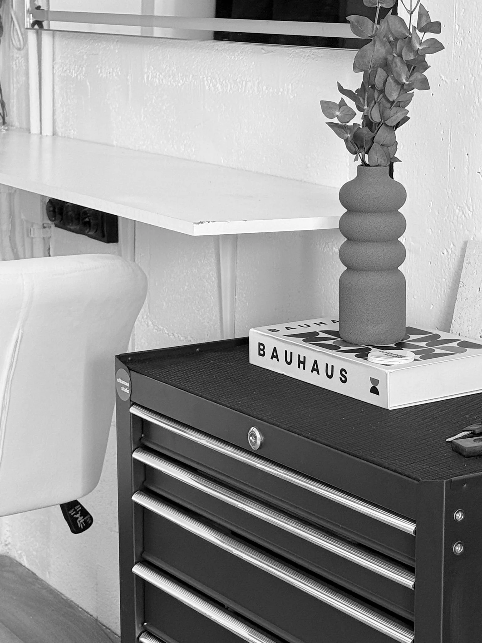 A black and white photograph showcases a room with a white wall, a white table, and a black rolling tool chest. On top of the tool chest is a rectangular box titled 'BAUHAUS' with a stylized black splash logo, and a decorative vase with a textured su
