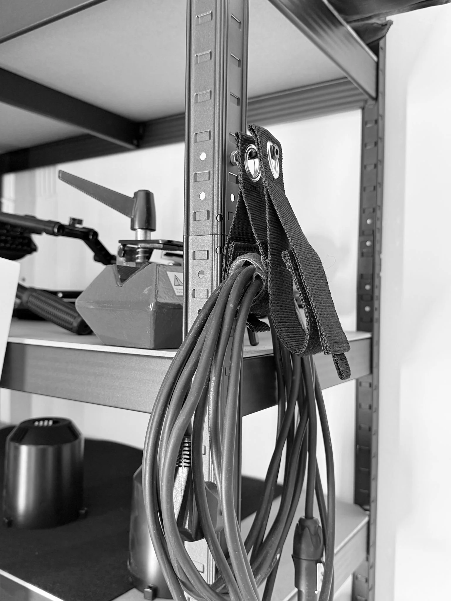 Black and white photo of a metal shelf with various cables hanging from hooks and a clamp attached to the side. The shelf is part of a storage or work area.