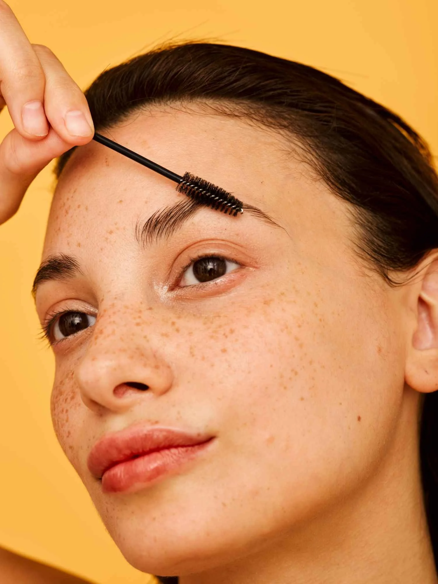 A close-up of a young woman with freckles applying mascara to her eyebrows against a yellow background.