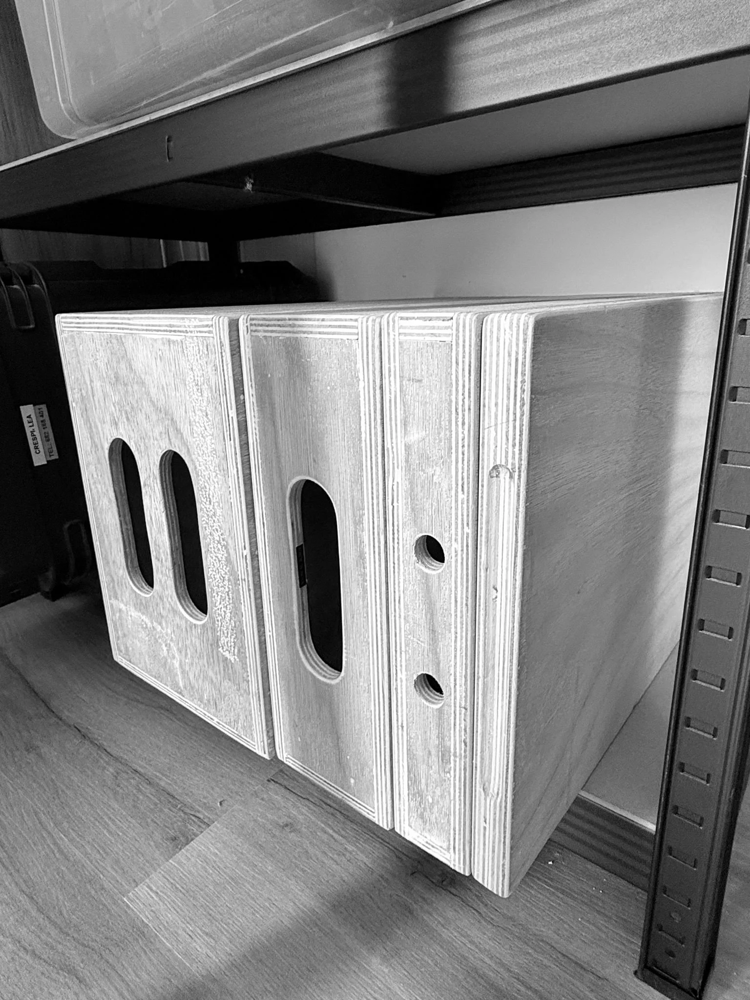 A wooden shelving unit with multiple oval-shaped cutouts in the front, placed on a wooden floor. Part of a gray plastic container and a black object are visible on top of the unit.