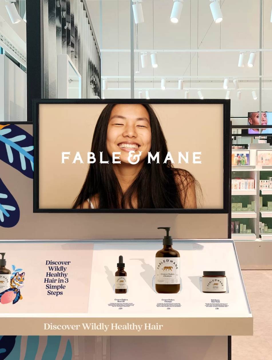 Display stand in a store showcasing Fable & Mane hair products, including a serum and a cream, with a large screen behind it showing a smiling woman with long dark hair and the Fable & Mane logo.