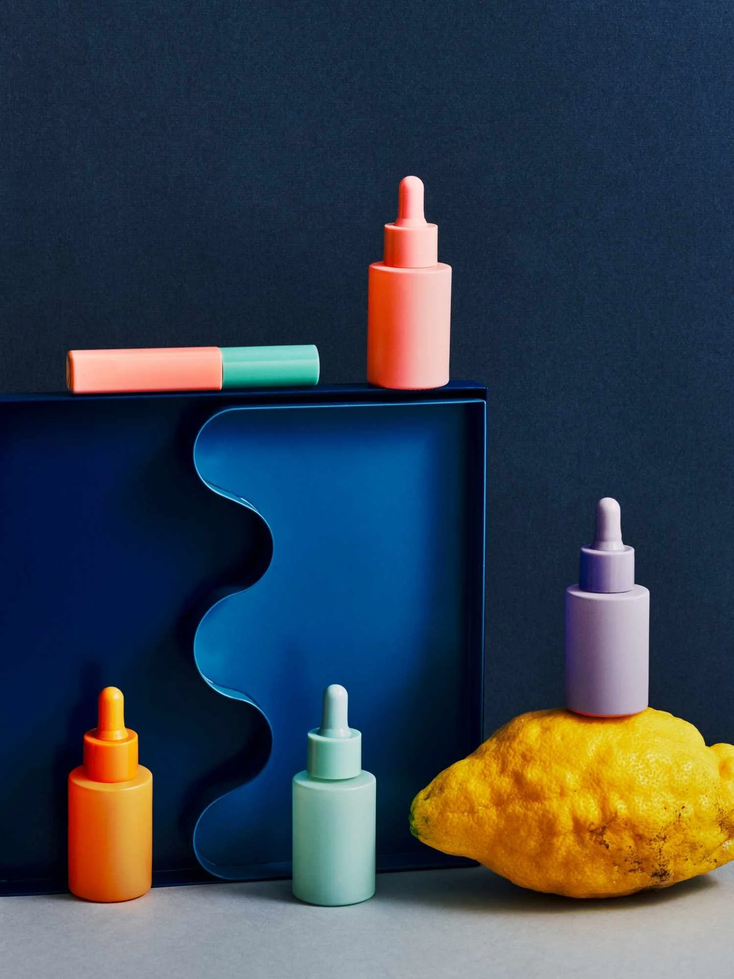 Colorful cosmetic bottles and droppers arranged next to a yellow textured object on a flat surface against a dark background.