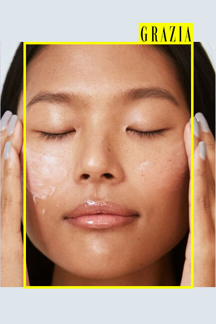 Close-up of a woman with clear skin and closed eyes, applying a skincare cream on her face, with the word 'GRAZIA' in yellow at the top right corner.