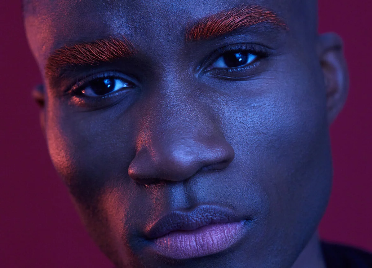 Close-up of a person's face with dark skin, highlighted by purple and pink lighting, showing detailed skin texture, dark eyes, natural lips, and eyebrows.