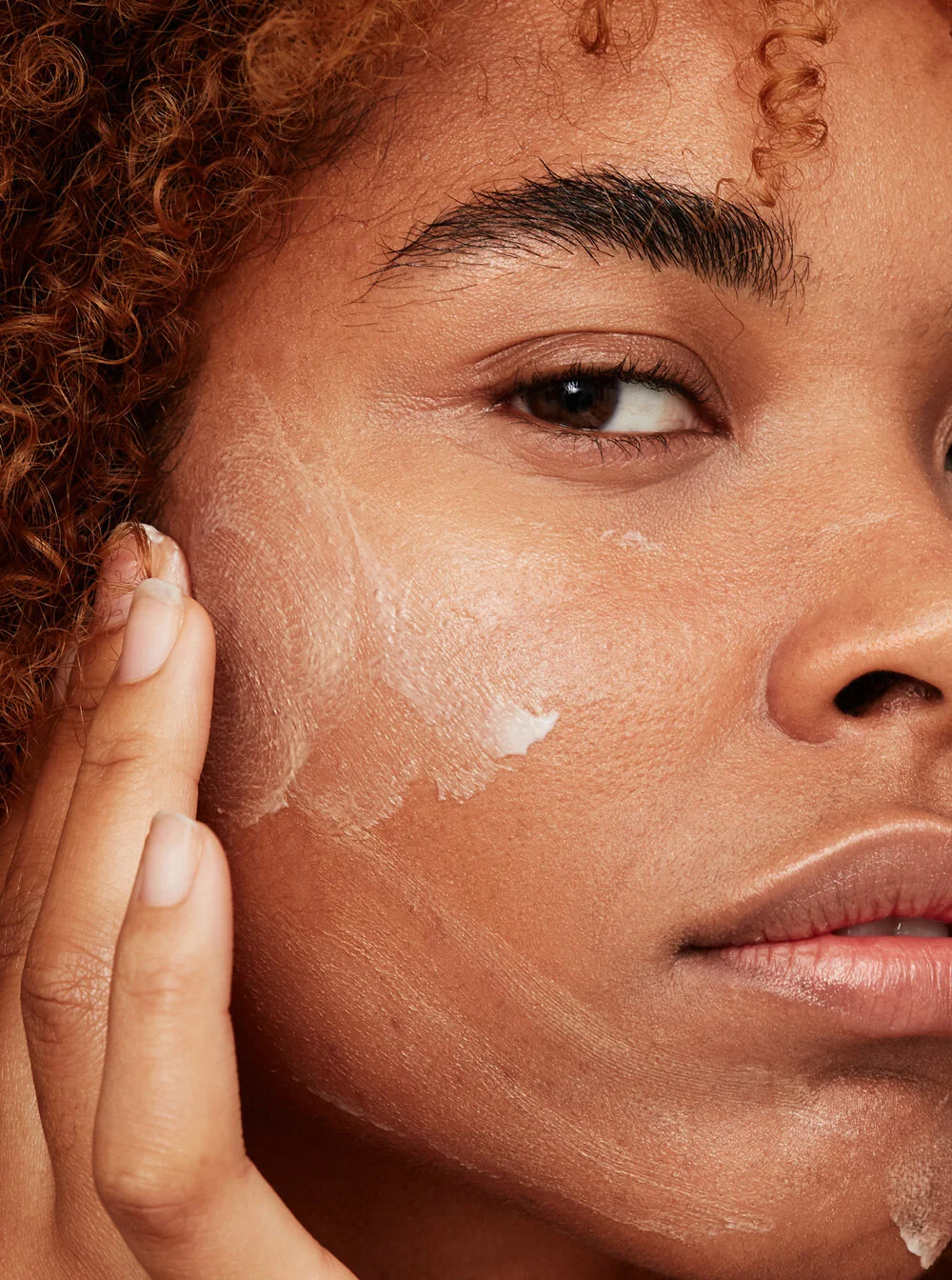 Close-up of a woman's face with a small amount of skincare or moisturizer applied to her cheek, with her fingers gently touching her skin.