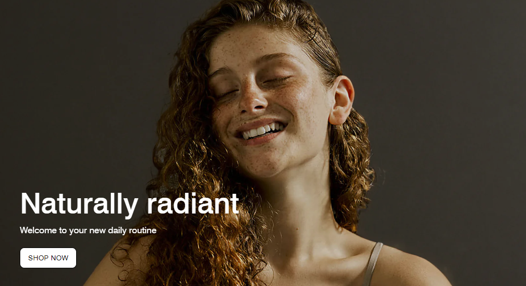 A woman with long, curly red hair smiling softly with her eyes closed, wearing a light-colored tank top, against a dark background.