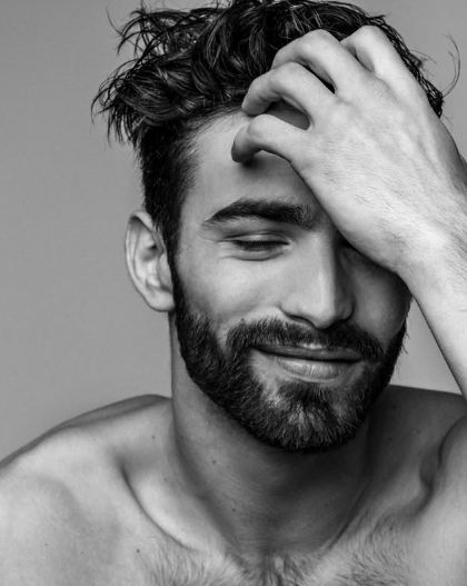 Black and white photo of a man with a beard and tousled hair, smiling with eyes closed, hand resting on his forehead.