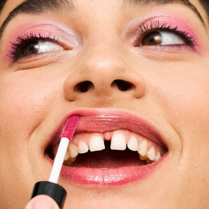 Close-up of a person's face applying pink lipstick, with pink eyeshadow and visible upper teeth.