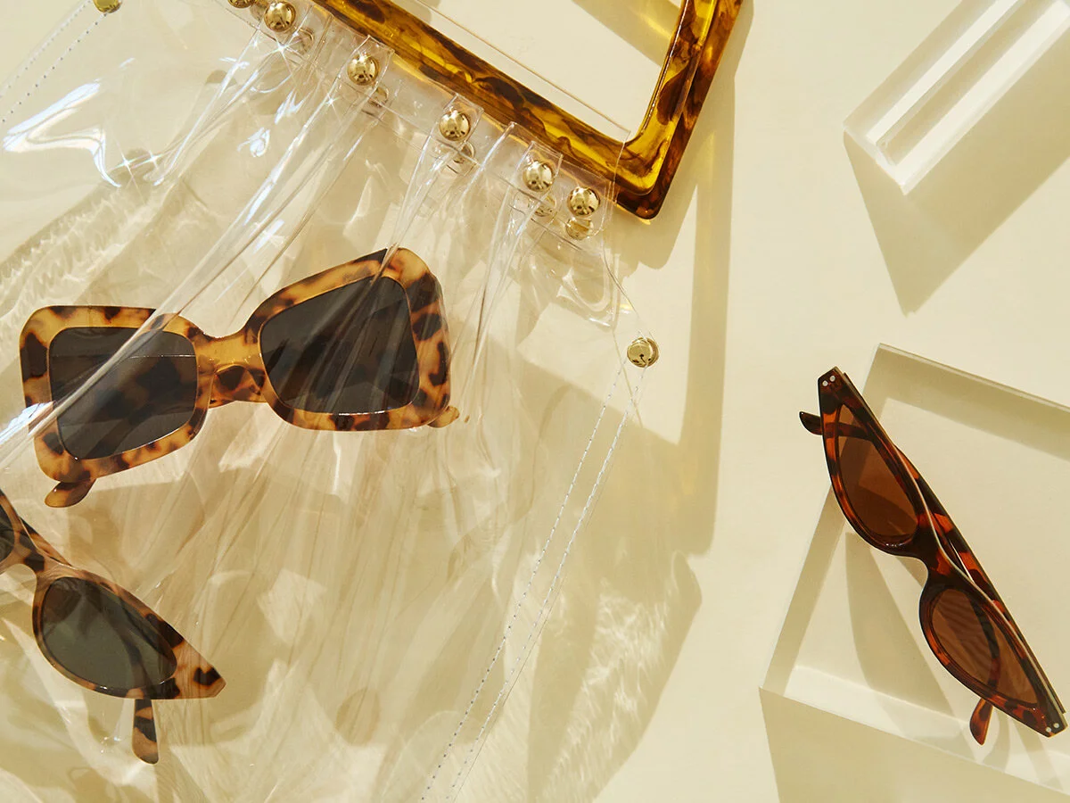 Two pairs of tortoiseshell sunglasses, one on a clear plastic sheet and one on a white wall, with a decorative box with gold ball details and small white shelves casting shadows in the background.