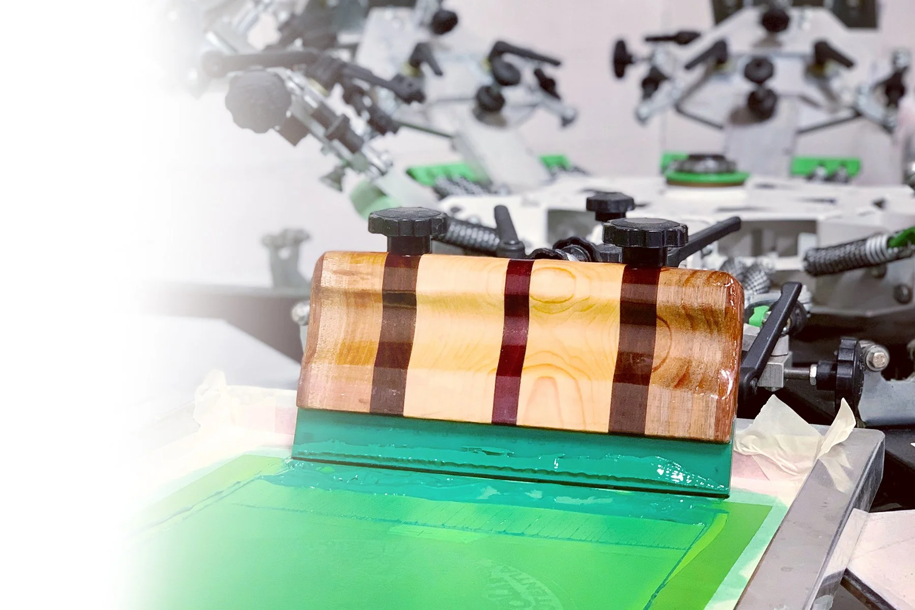 A custom wooden screen printing squeegee sitting inside of a screen with green ink and a printing press in the background.