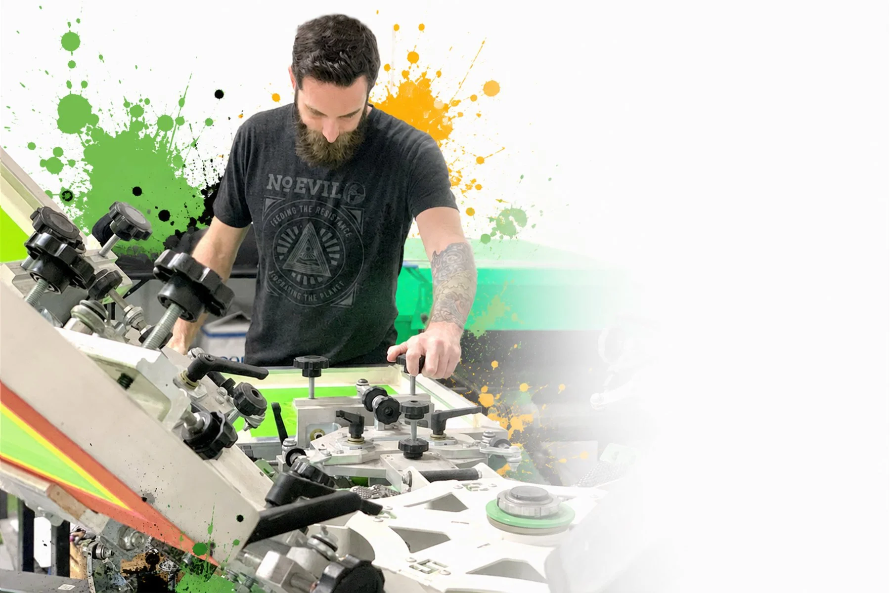 A man with a beard working on a manual screen printing press with colorful paint splashes in the background.