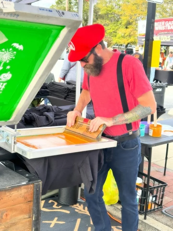 A man with a beard wearing a red shirt, black suspenders, and a red cap printing t-shirts at an outdoor event. He is using squeegee to push ink through a screen. There are tables with supplies and other people in the background.