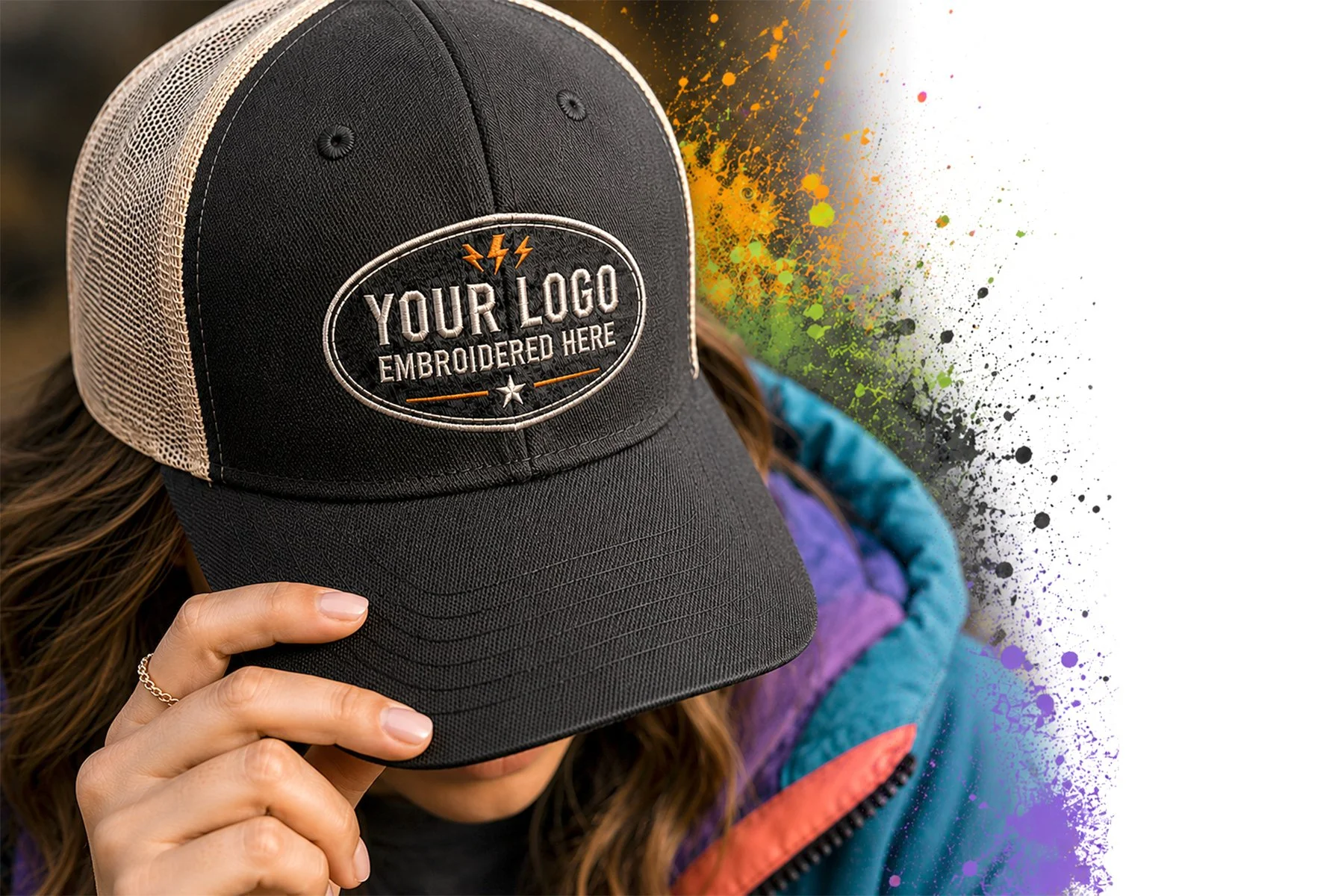 Person wearing a black trucker hat with an embroidered patch that reads "Your Logo Embroidered Here" and is holding the hat's brim with their right hand. The background features colorful paint splashes.