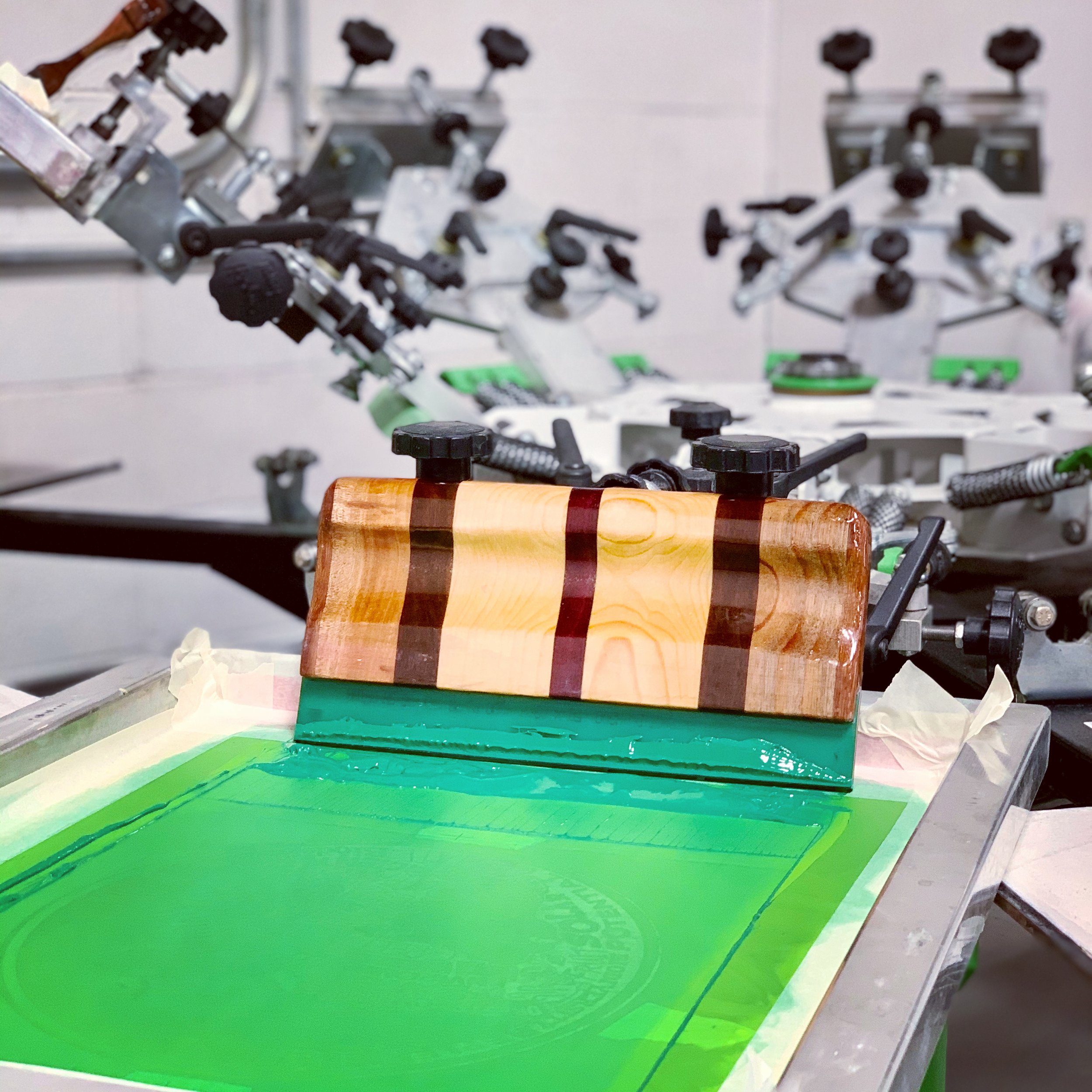 Custom hand crafted wooden screen printing squeegee sitting inside a screen set up on a printing press flooded with green ink.