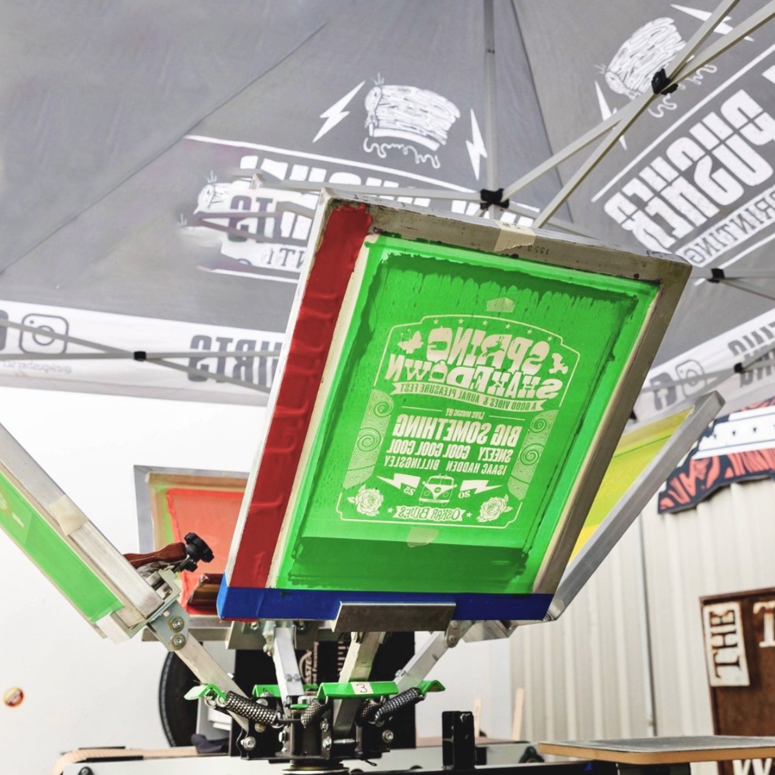 Close-up of a screen coated with green emulsion attached to a 4 color printing press inside of an event tent.