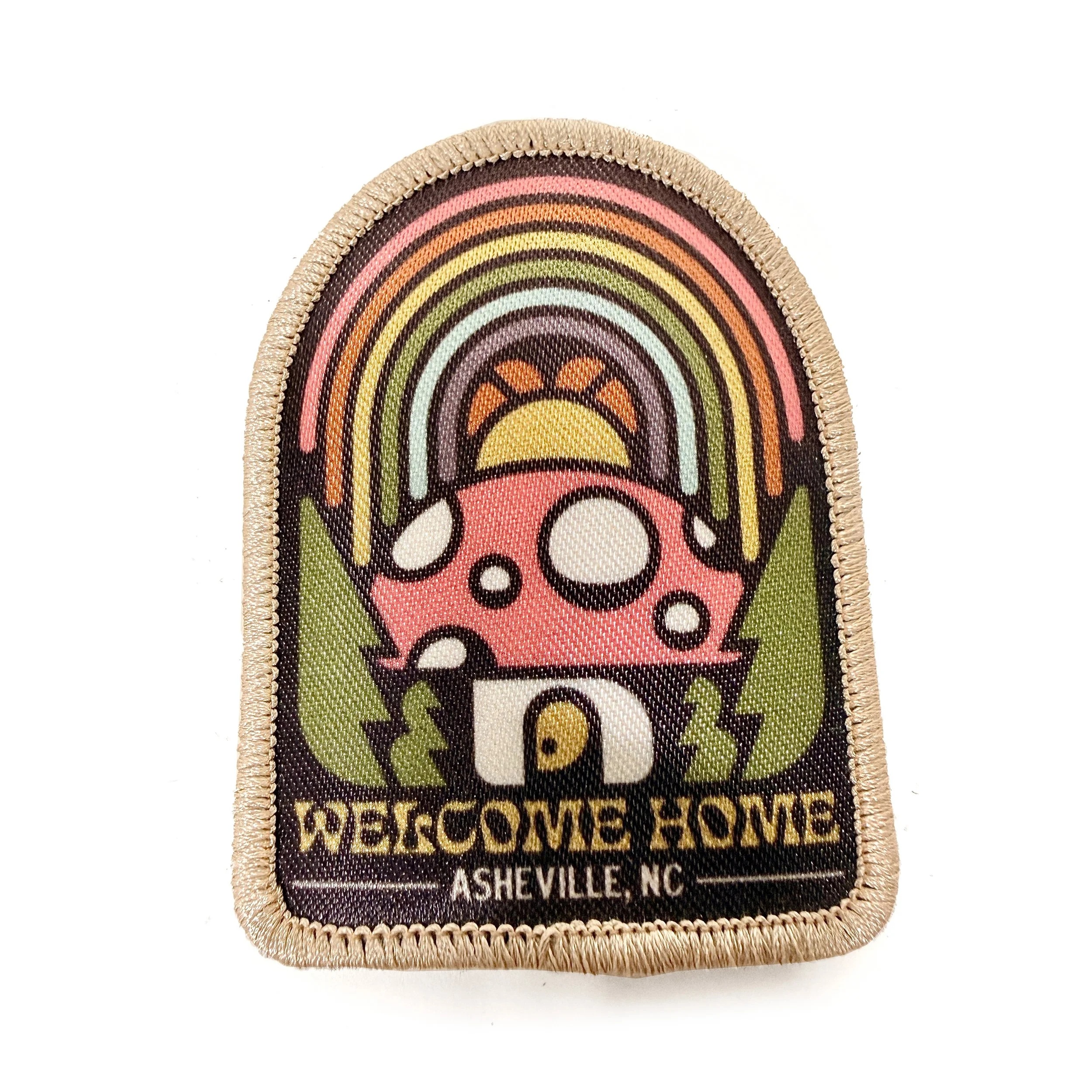 Close-up of a colorful embroidered patch with a rainbow and a skull, bordered by beige stitching and the words "WELCOME HOME" and "ASHEVILLE NC" at the bottom.