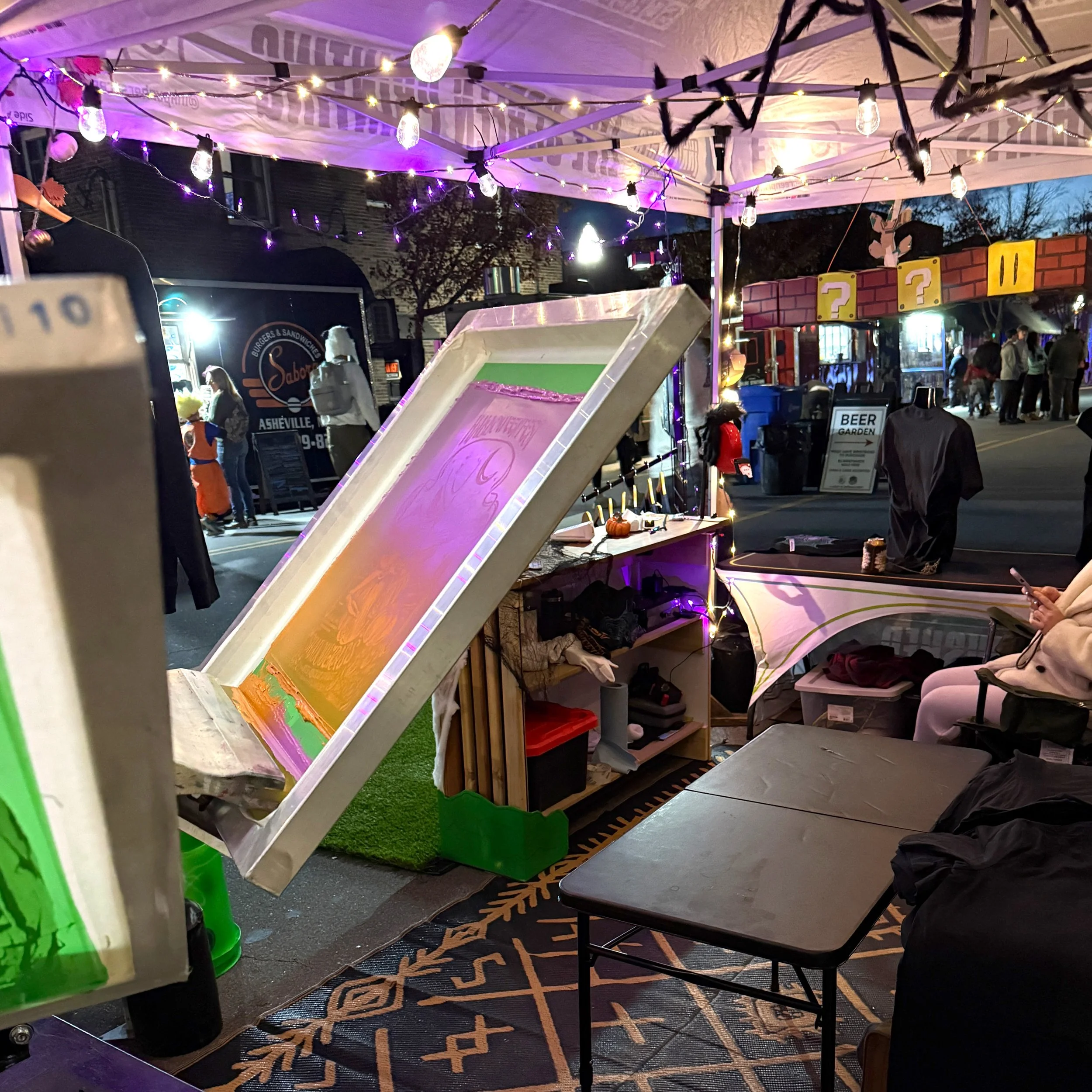 Nighttime scene at a street festival showing the inside of a screen printing booth.