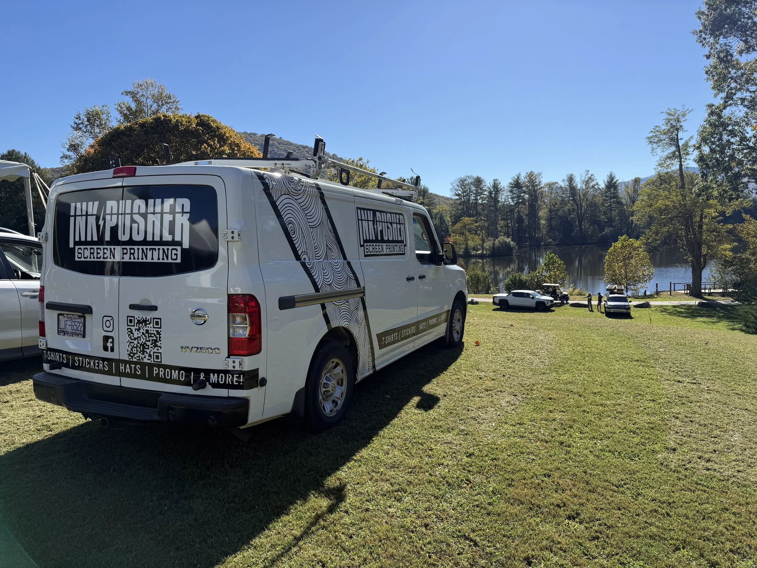 A white van with “Ink Pusher Screen Printing” logo and contact information parked on a grassy area near a lake and trees, under a clear blue sky.