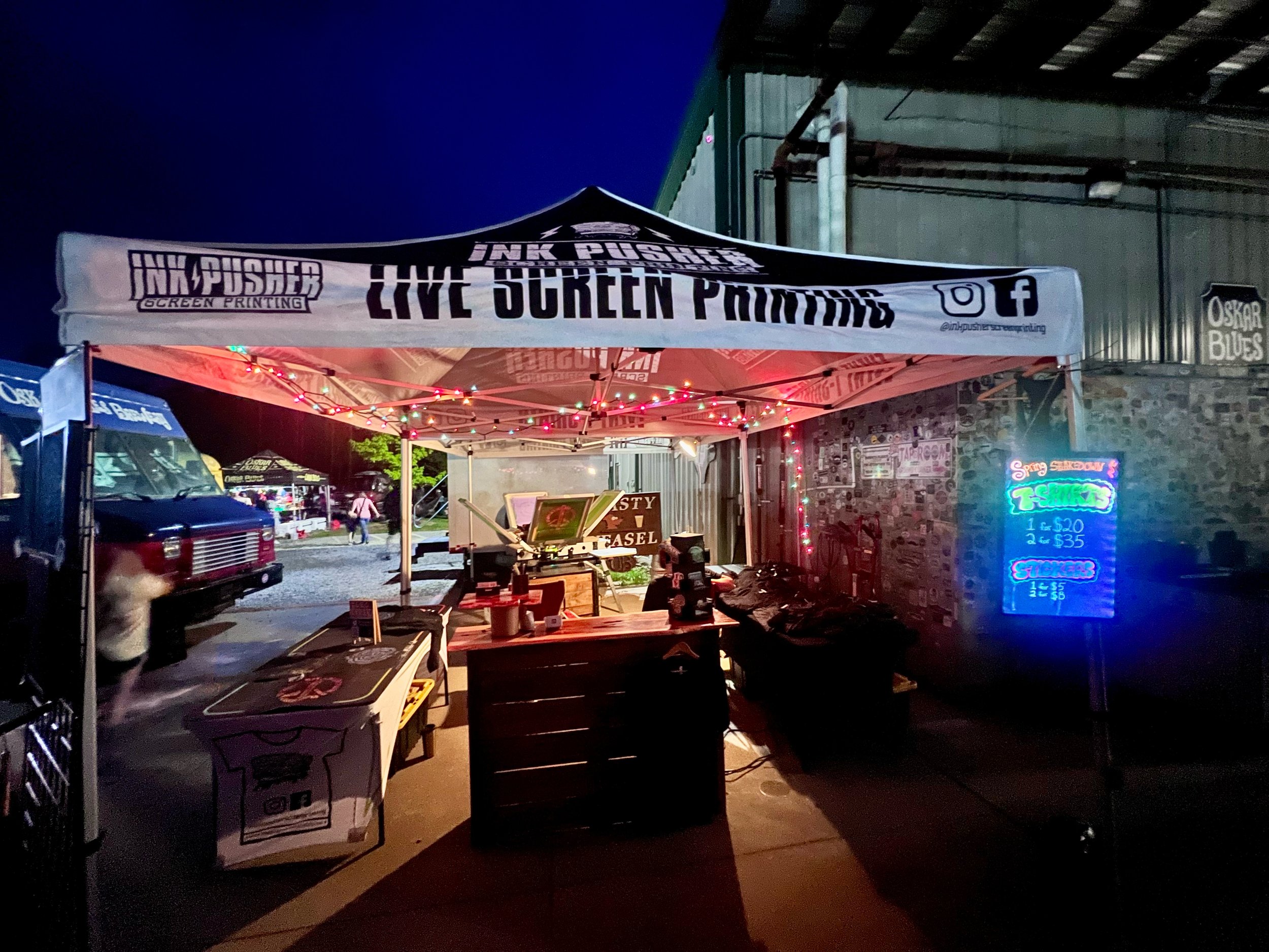 A night view of a live screen printing booth with a canopy reading 'INK PUSHER SCREEN PRINTING' and social media icons. The stall has colorful string lights, and a neon sign offers pricing for stickers.
