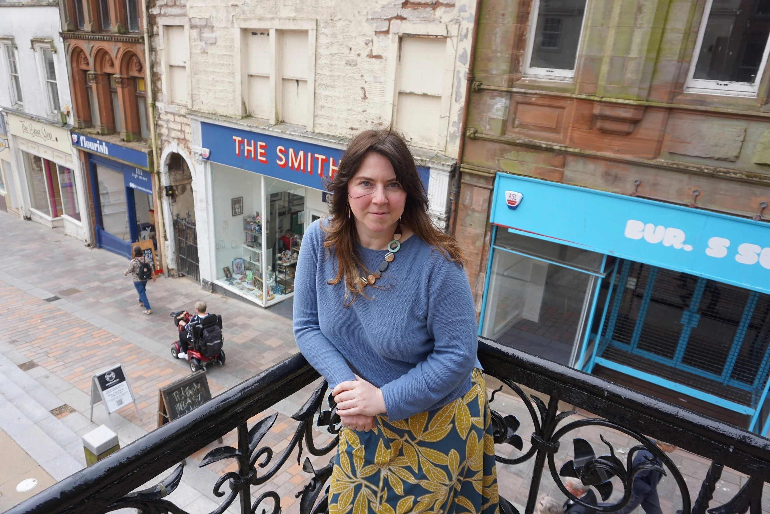 Heather Taylor leans on the railings of the Midsteeple building. Behind her are MSQ buildings on the high street.