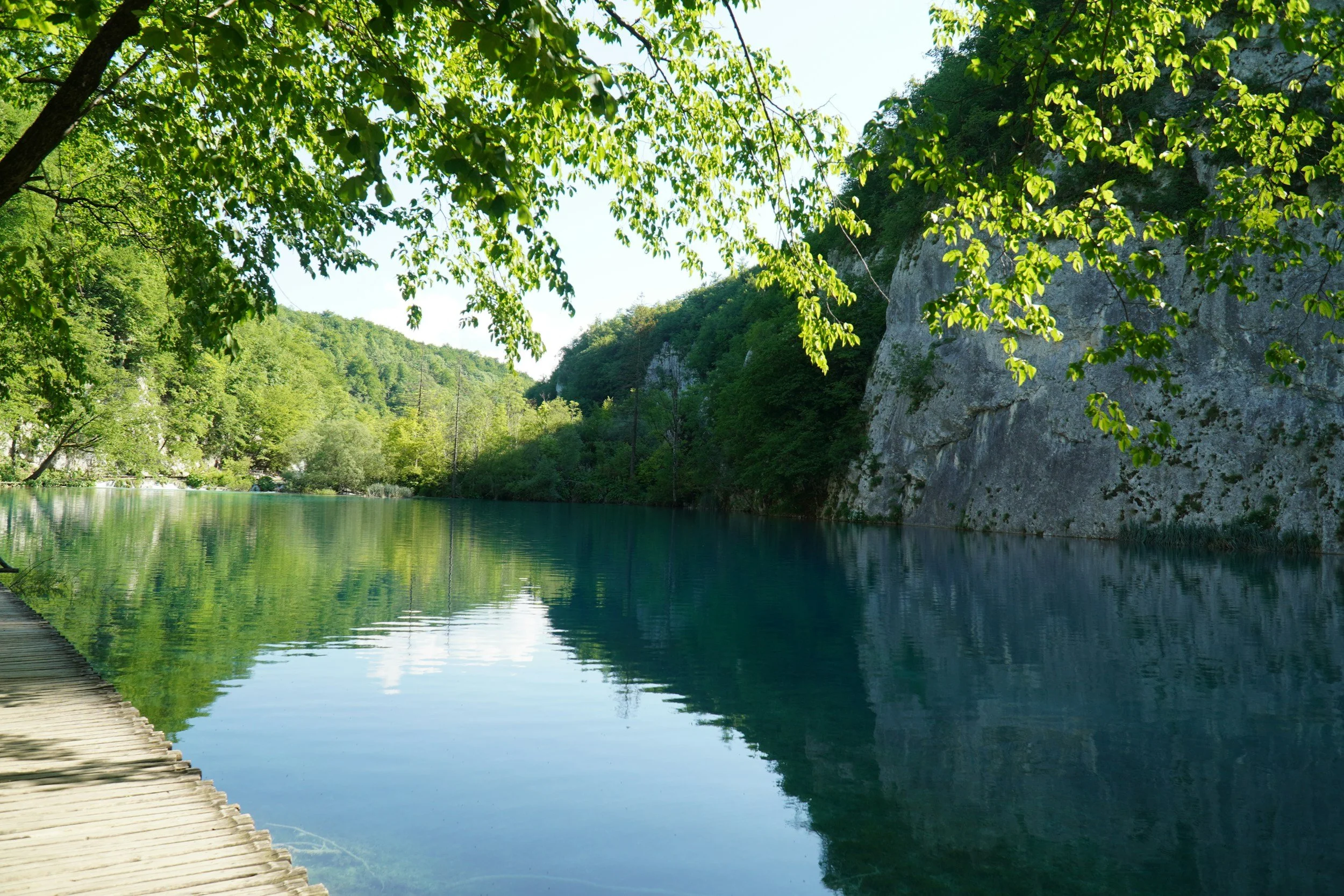 A scenic view of a river surrounded by lush green trees and a rocky hillside, with a wooden pathway on the left side of the image.